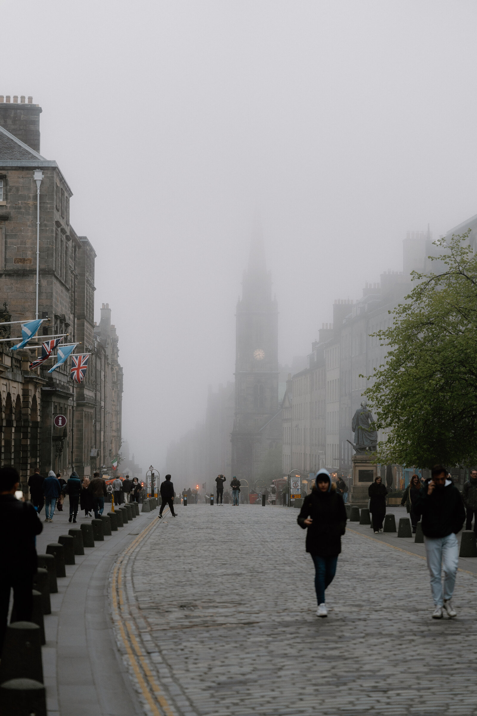 Pedestrians walk a misty Royal Mile beneath flags, the Hub's spire fading into fog ahead.