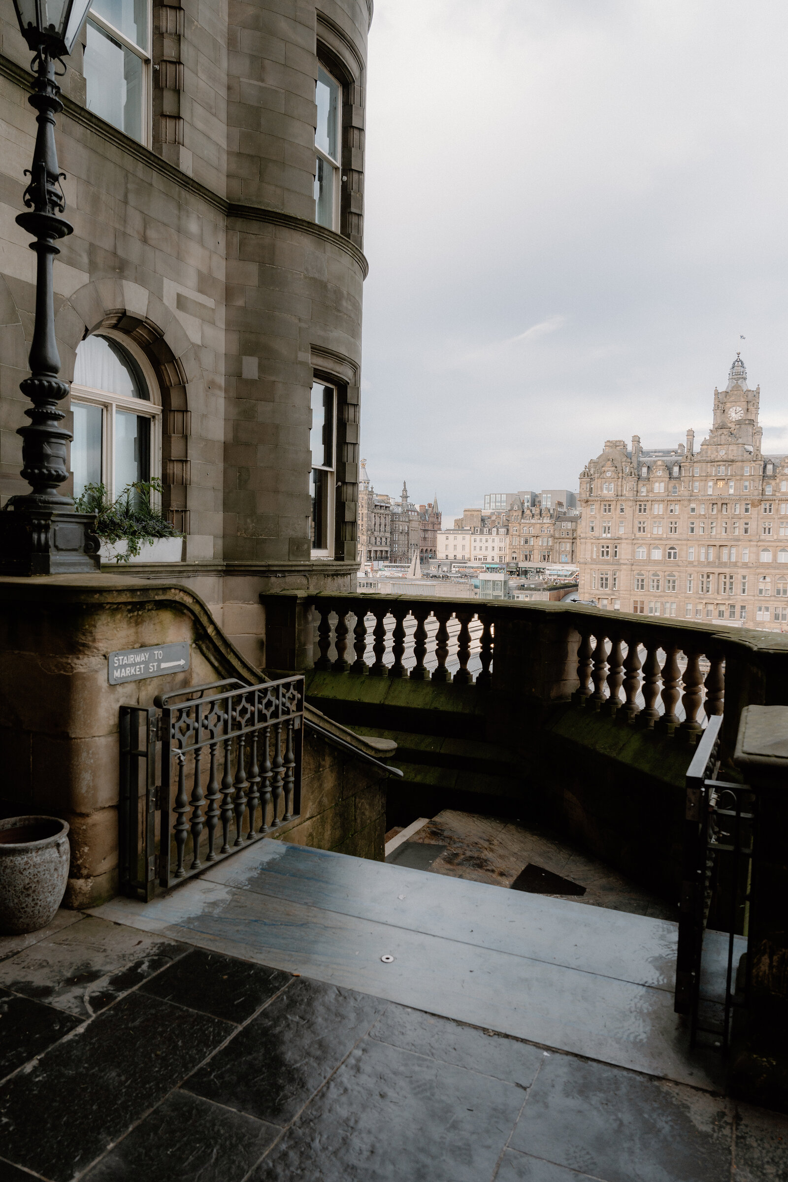A stone balcony overlooks Edinburgh's rooftops toward the Balmoral Hotel's clock tower, a 'Stairway to Market St' sign on the railing.