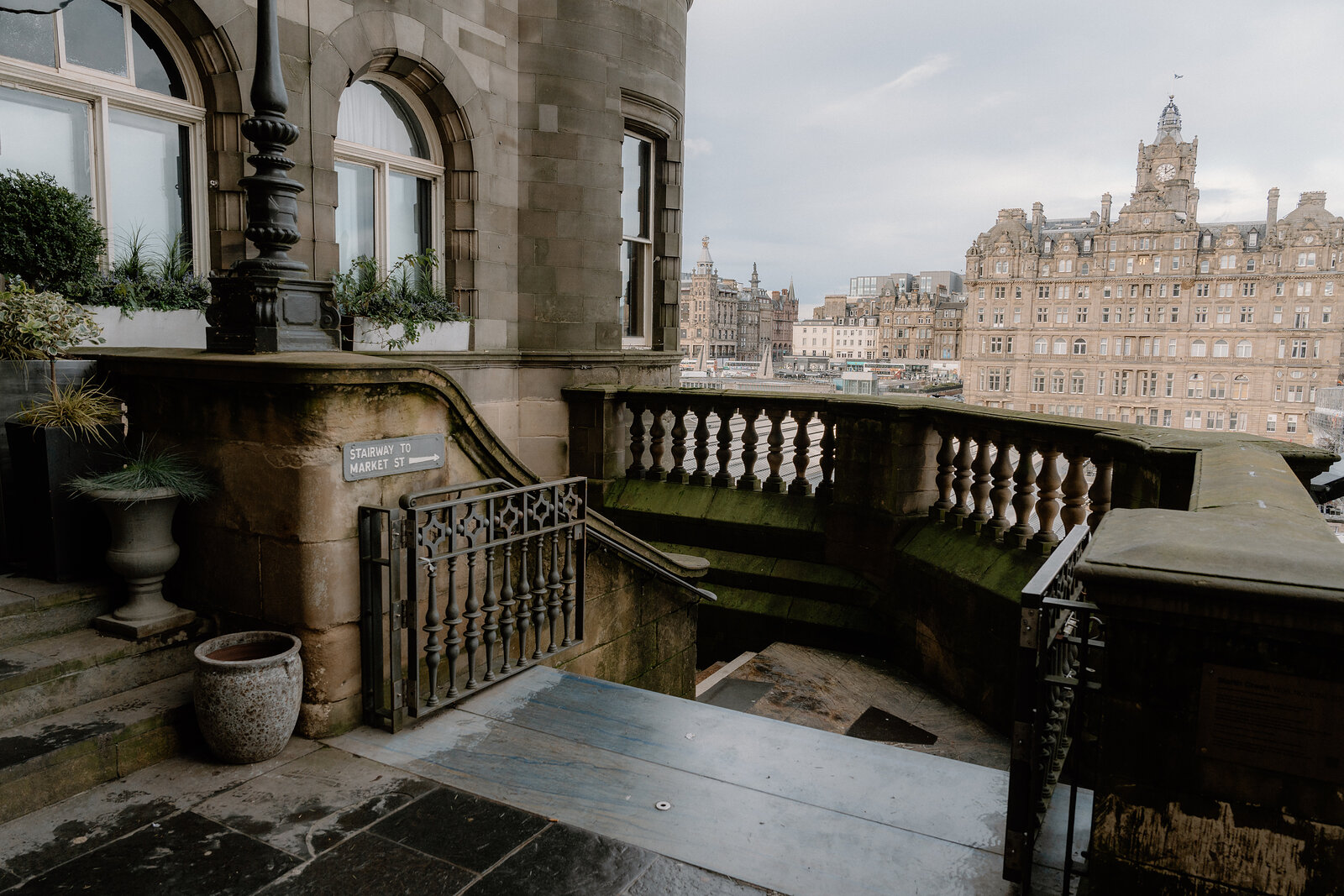 A stone balcony with urn planters overlooks Edinburgh's rooftops toward the Balmoral Hotel.