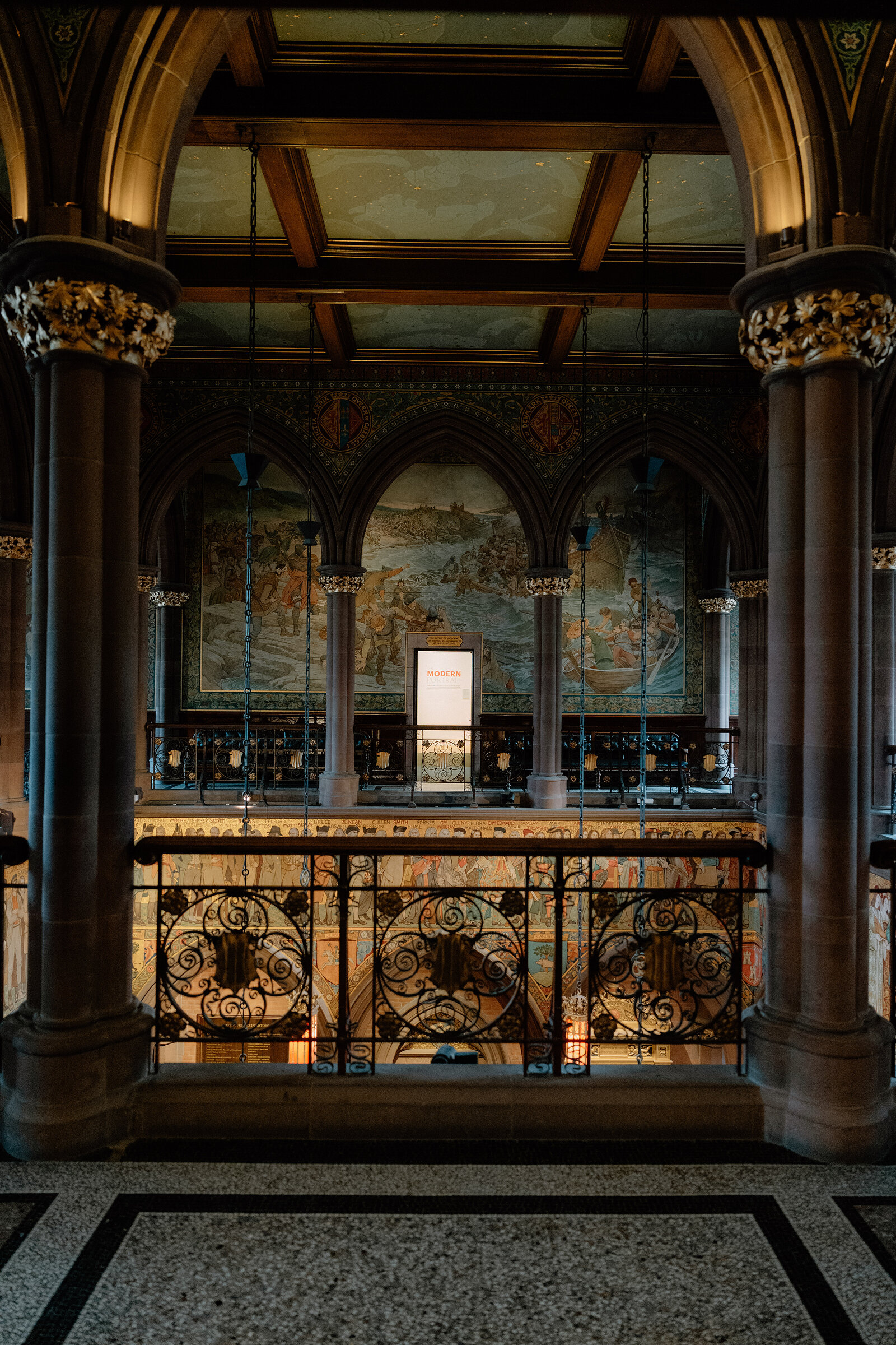 A painted processional frieze lines the upper gallery of a grand hall, wrought-iron railings below.