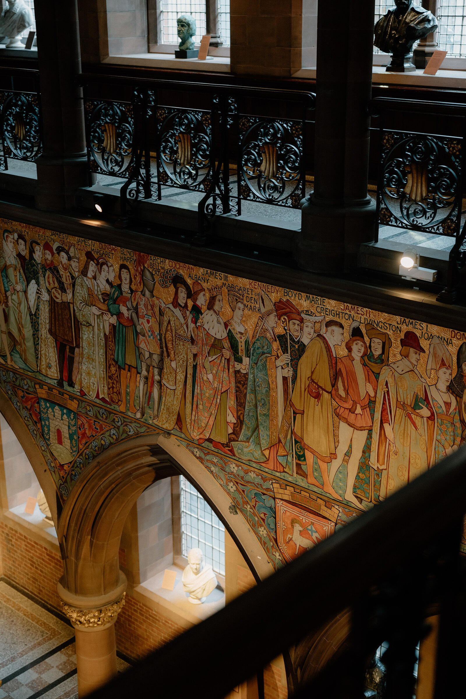 A painted frieze of historical figures runs above a stone archway in a grand hall, sculpted busts set below.