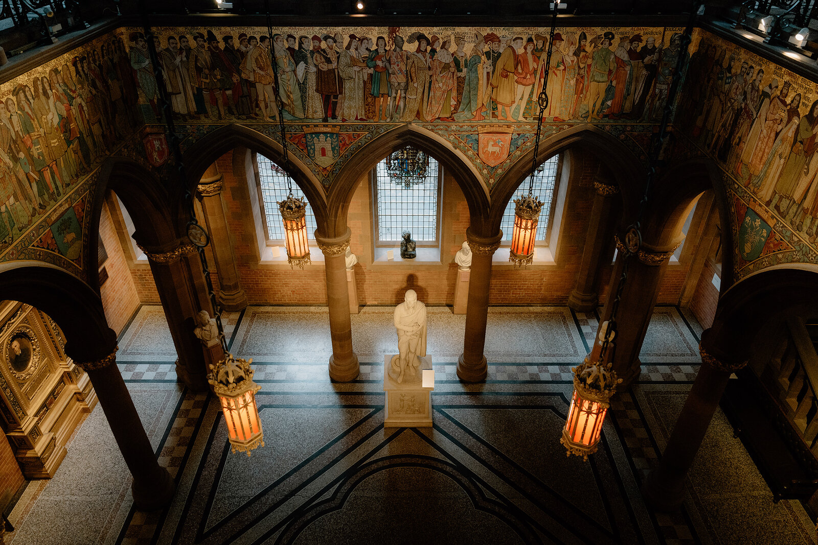 A marble statue stands beneath a painted frieze and hanging lanterns in a grand galleried hall.