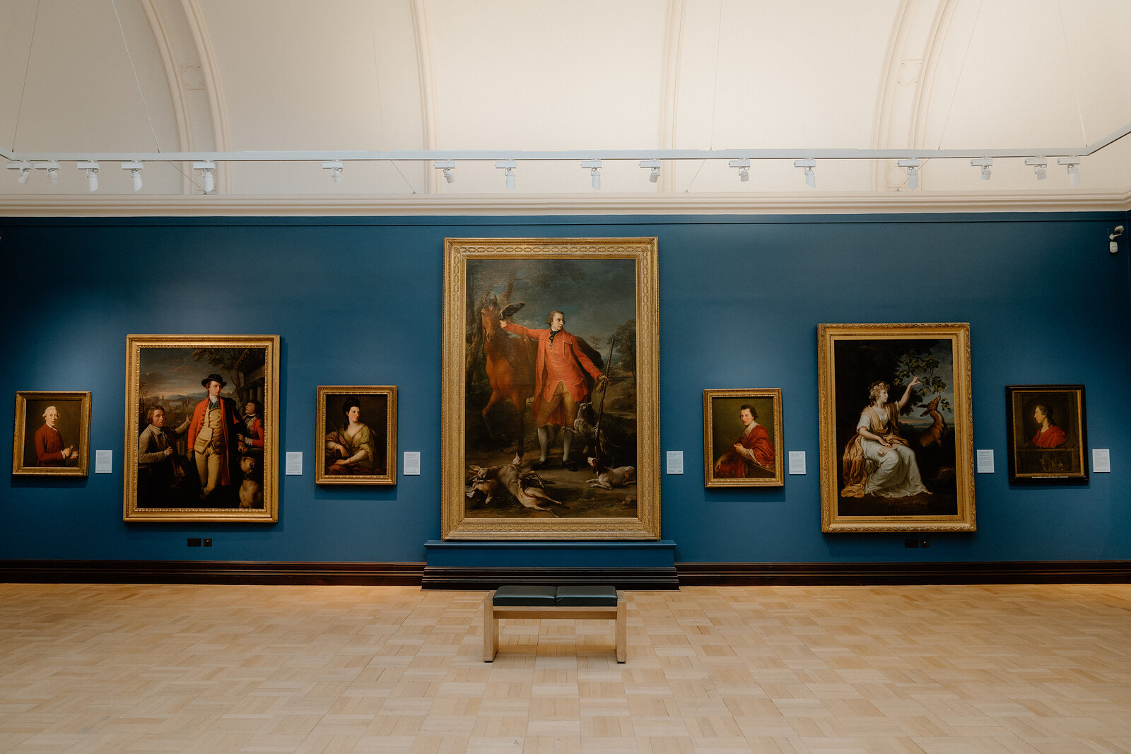 Gilt-framed portraits hang on a deep blue wall in the Scottish National Portrait Gallery, a bench set before them.