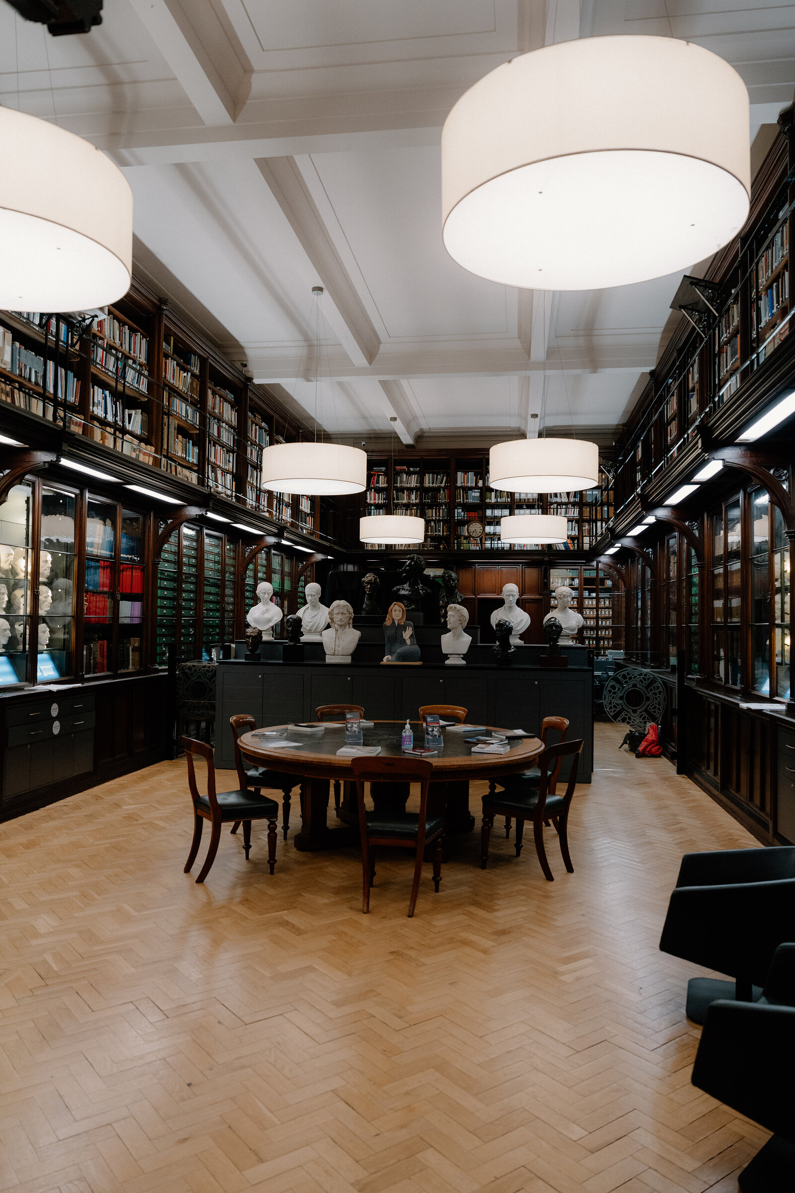 Marble busts sit on a round table beneath pendant lights in a wood-panelled library.