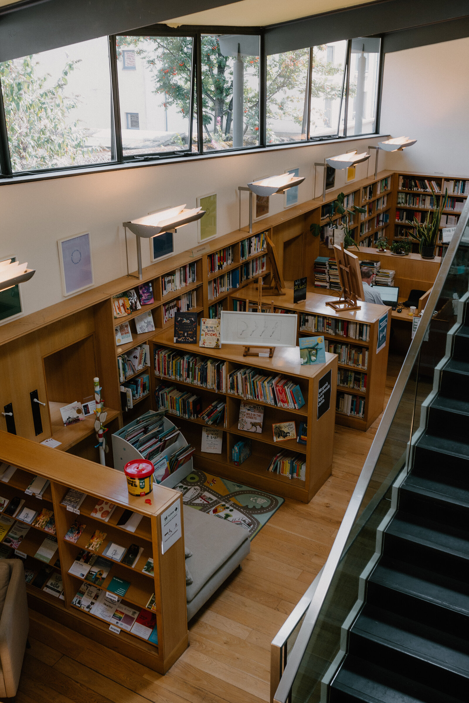 A children's library with wooden shelving and reading nooks sits beneath tall windows and a mezzanine staircase.