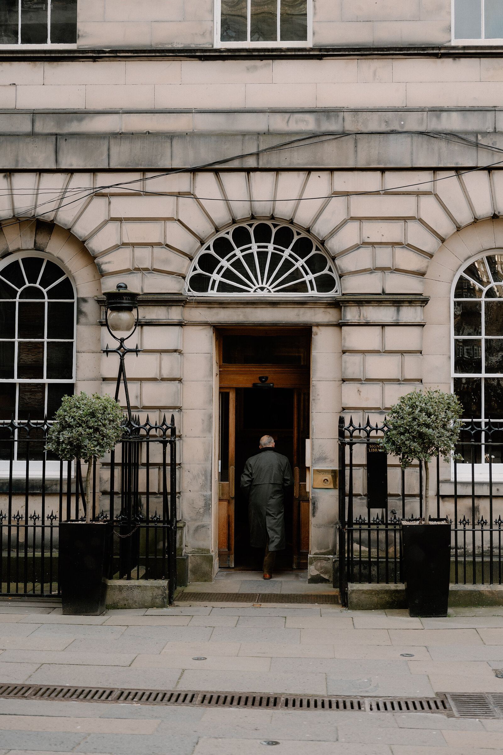 A man walks through a grand stone doorway flanked by potted trees and wrought-iron railings.