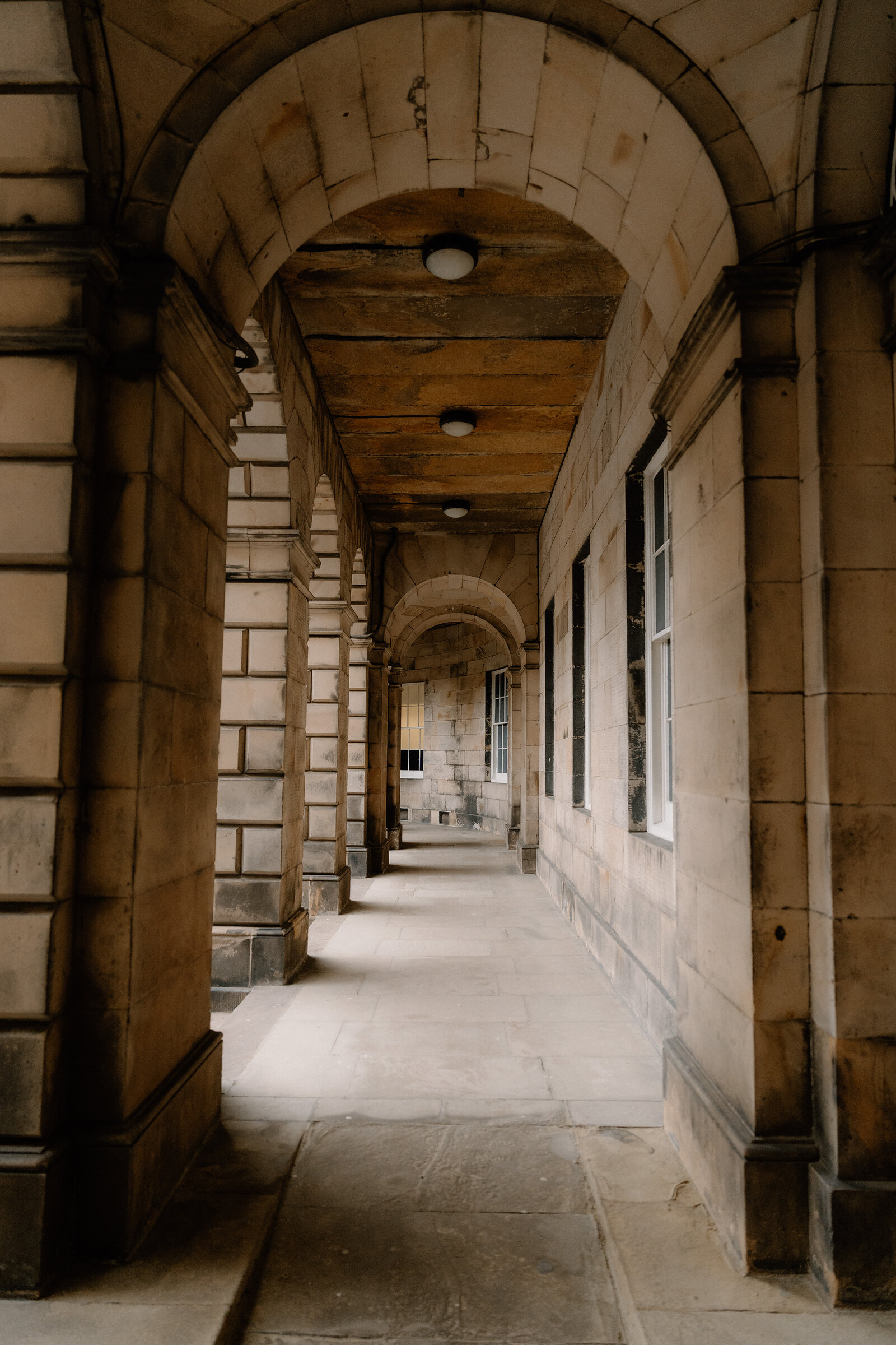 A stone colonnade lit by hanging lamps stretches beneath an arched ceiling in a grand building.
