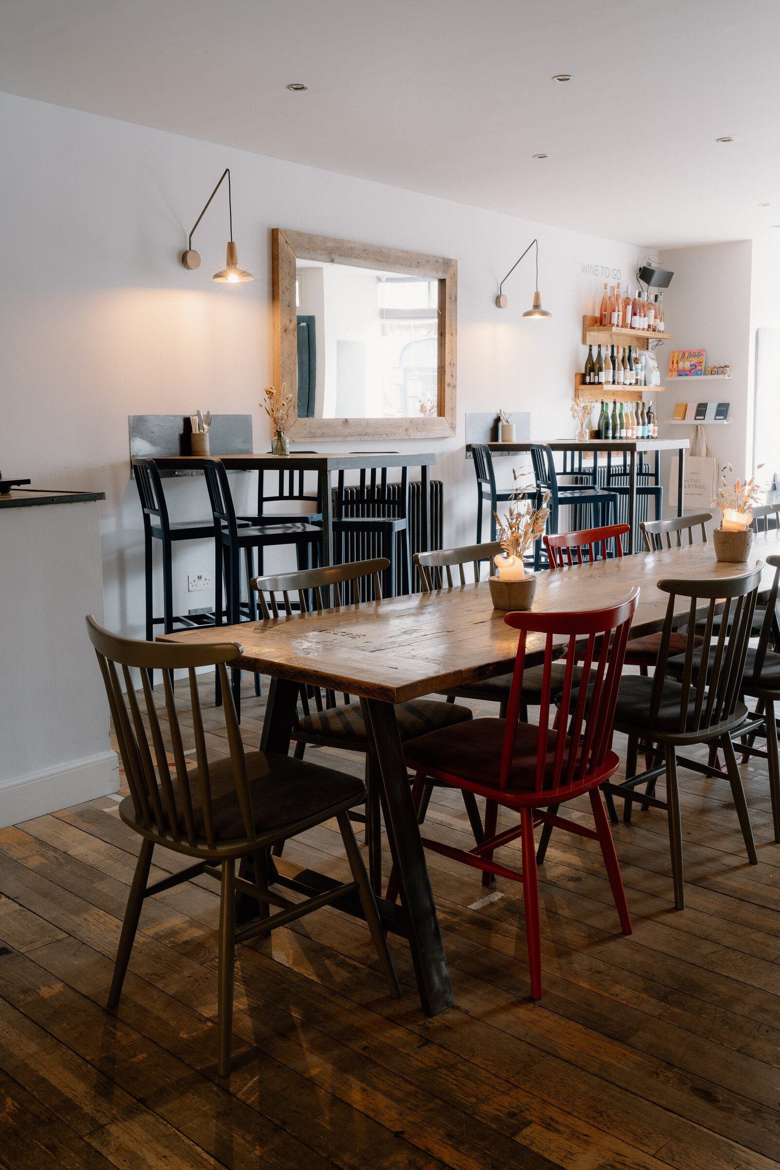 Mismatched wooden chairs surround a long table before a mirror and shelved bar in a restaurant.