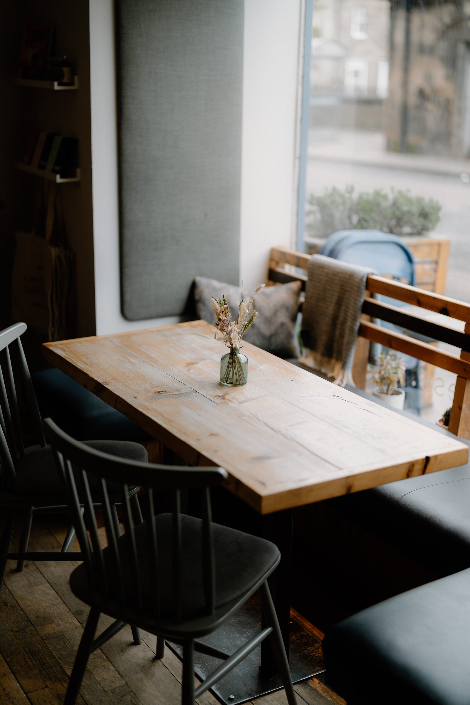 A wooden table with a small flower vase sits before a banquette in a cafe window.
