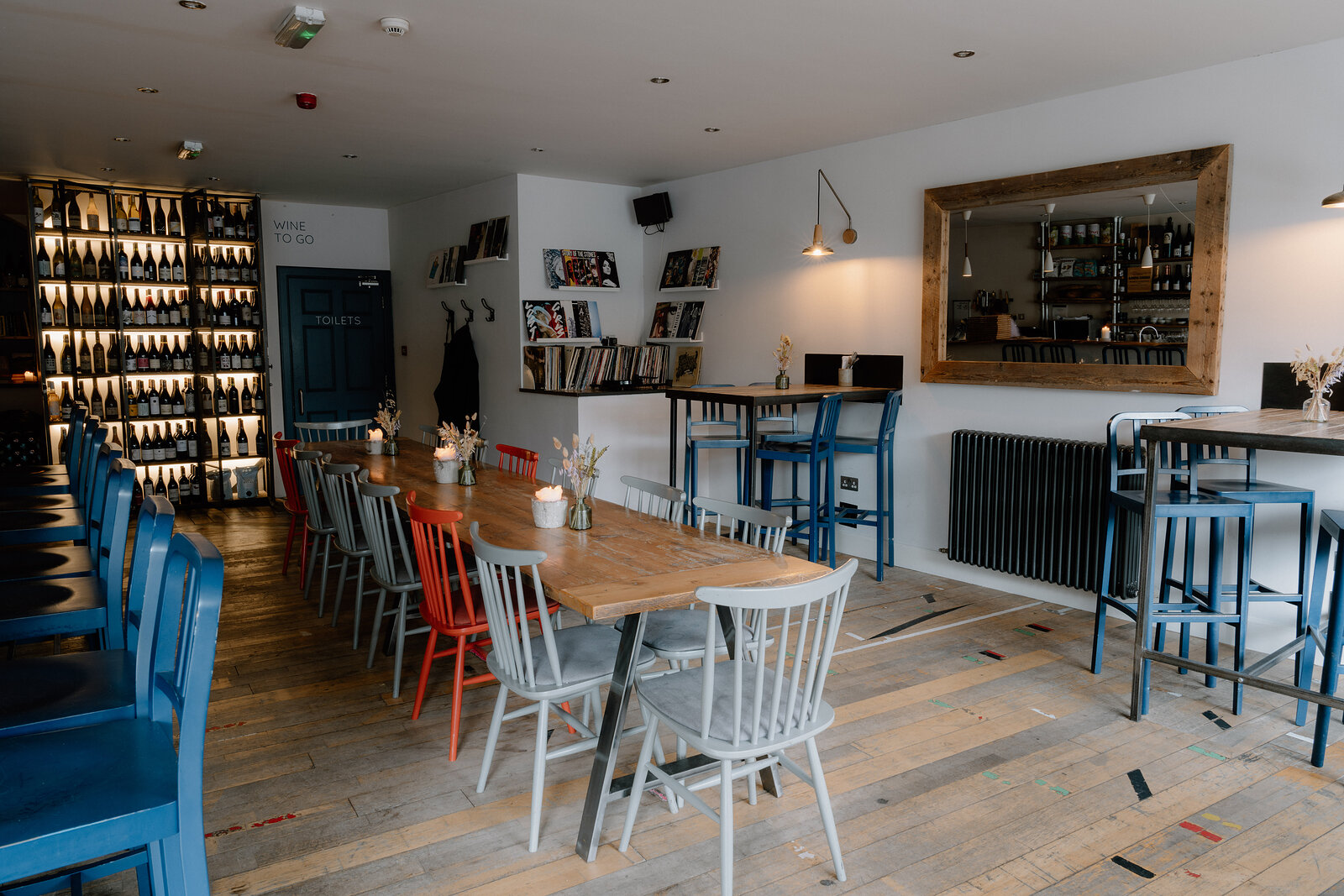 Wine racks line a wall beside blue and patterned chairs at wooden tables in a wine bar.