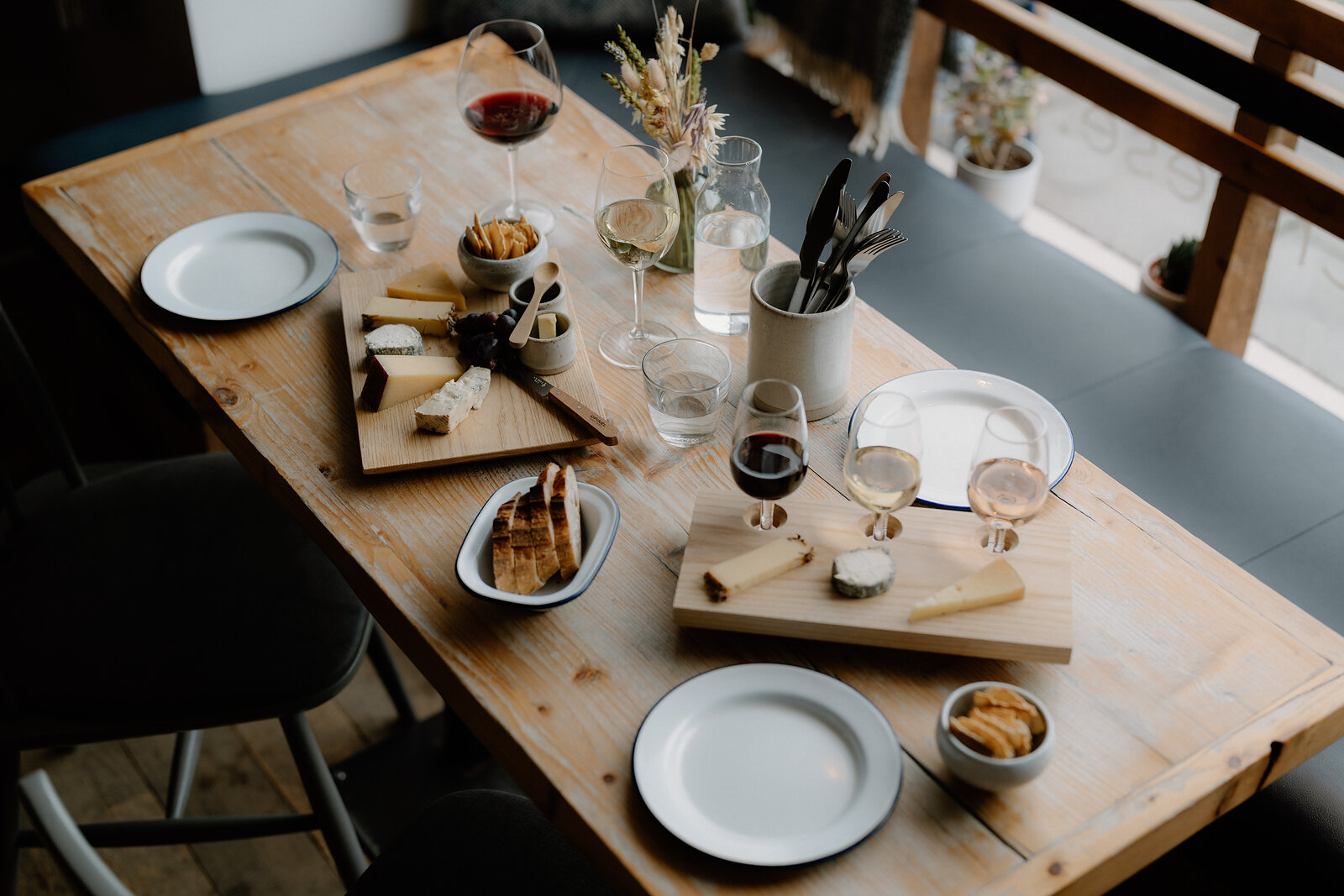 A cheese board and bowl of crackers sit beside glasses of wine and a small potted plant.