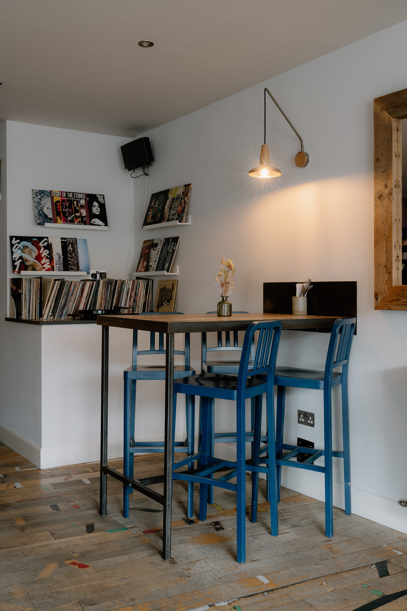 Blue stools sit at a high table beneath a wall-mounted lamp and shelves of vinyl records.