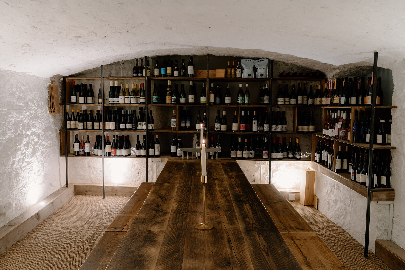 A vaulted stone wine cellar holds shelves of bottles along both walls, a long wooden table set with a candle down the centre.