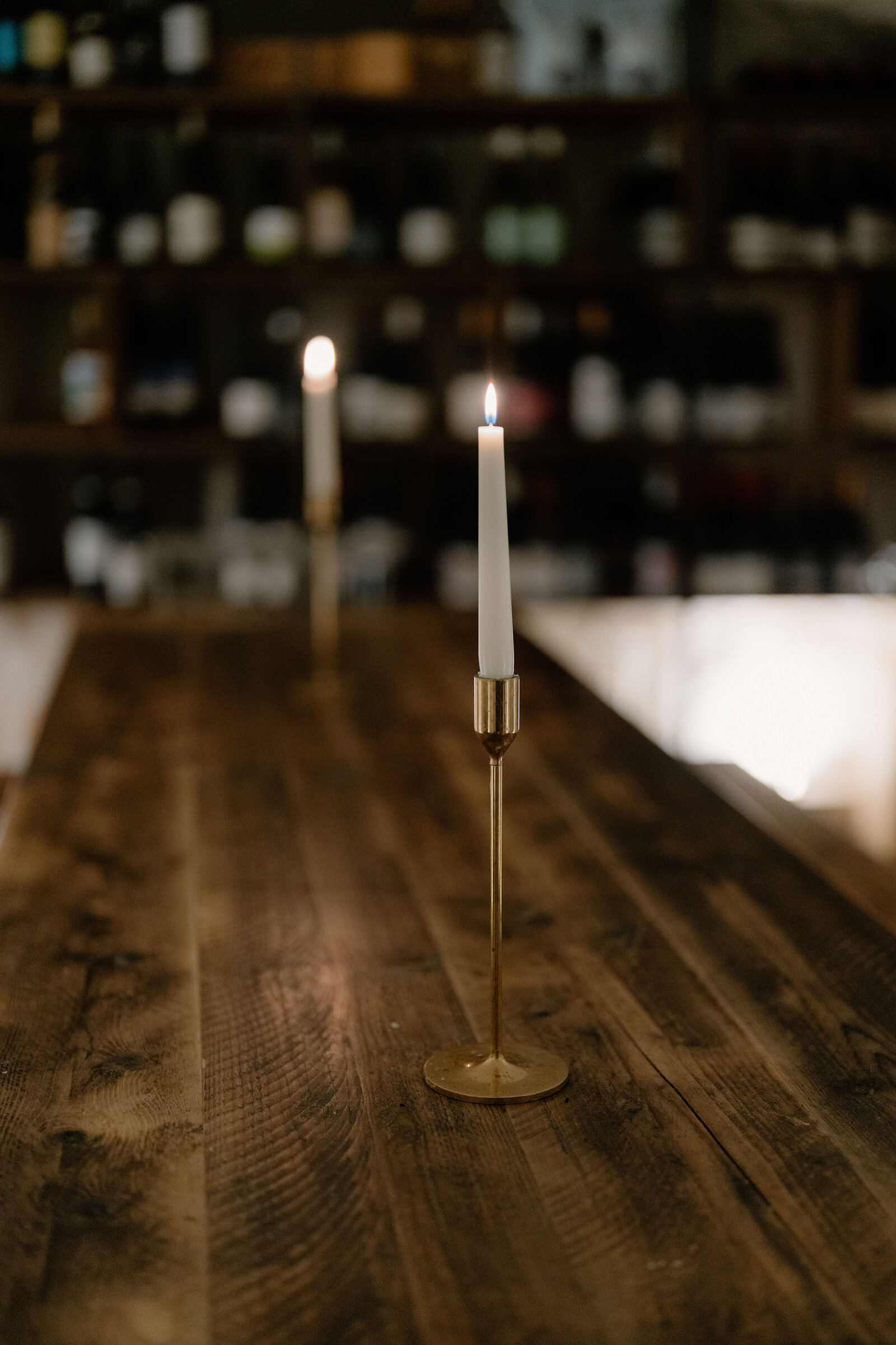 Lit candles stand on a wooden table before shelves of wine bottles in a dim cellar.