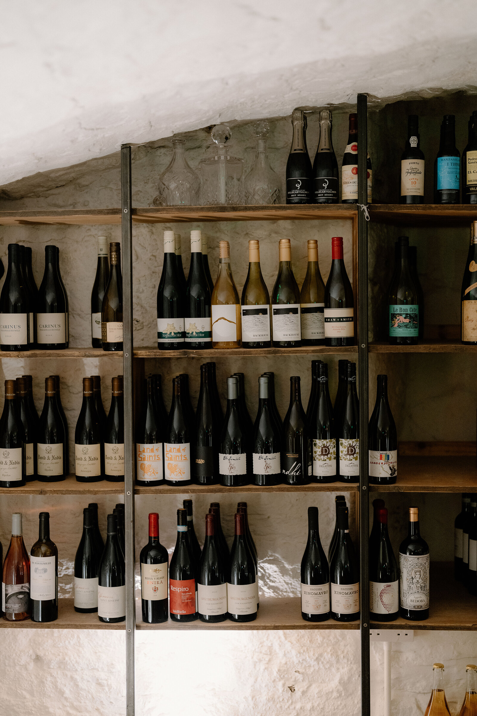 Wine bottles and glass decanters fill wooden shelves set into a vaulted stone wall.