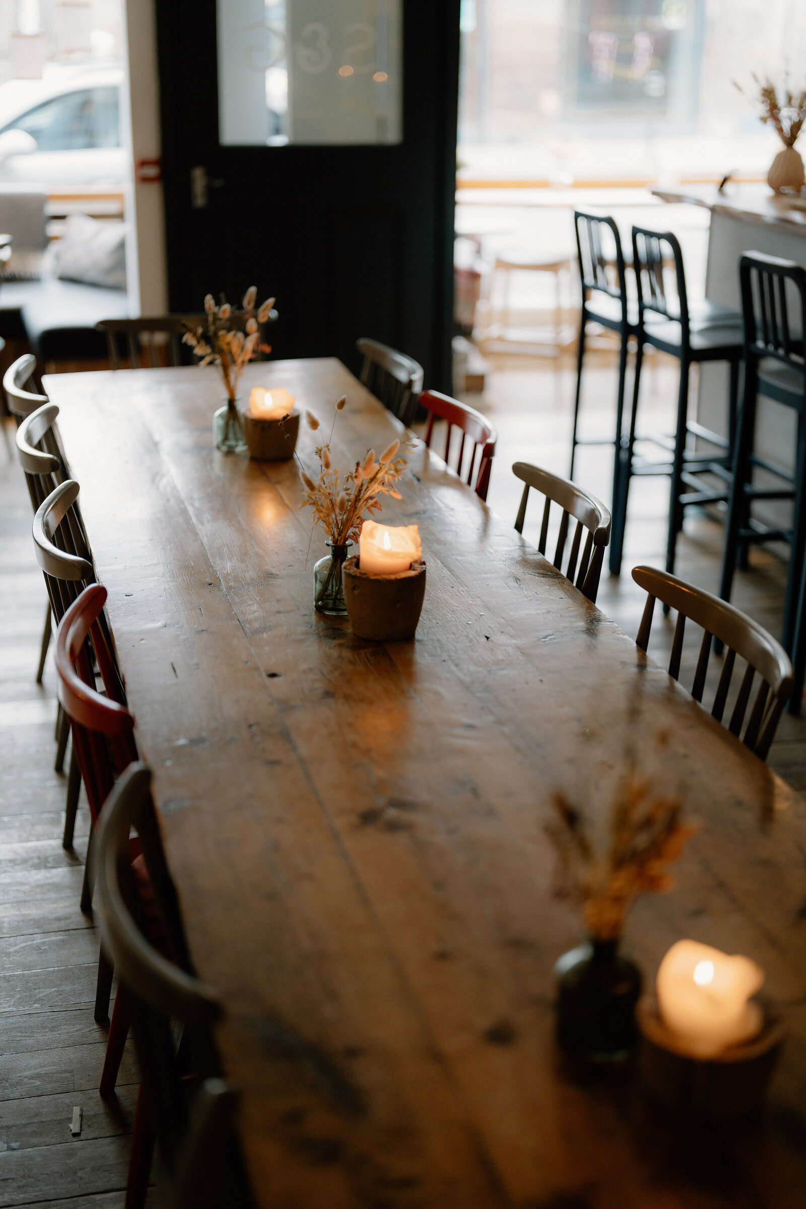 A long wooden table set with candles and dried flowers sits before a window and bar stools.