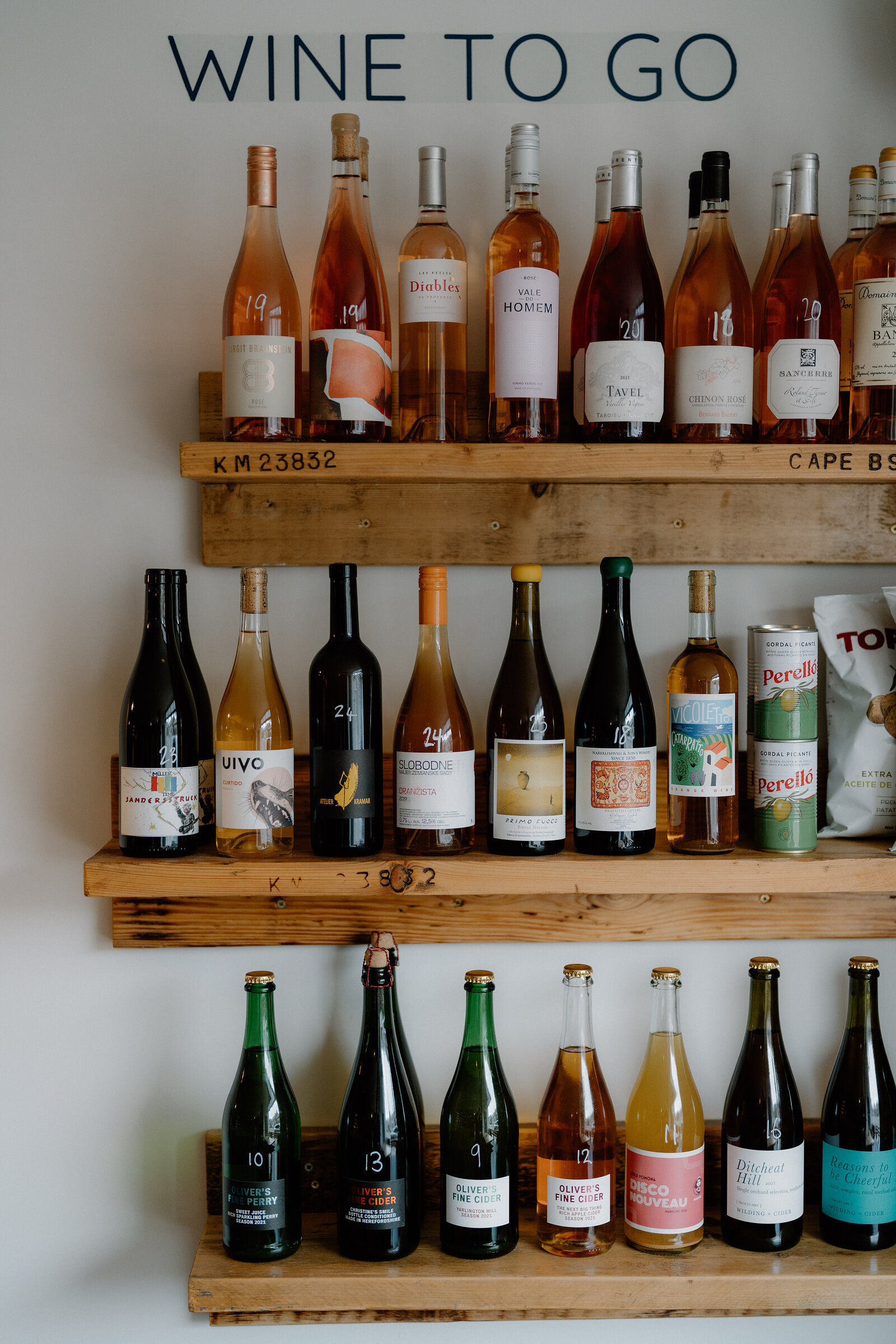Bottles of rosé, white and sparkling wine line wooden shelves beneath a sign reading 'Wine To Go'.