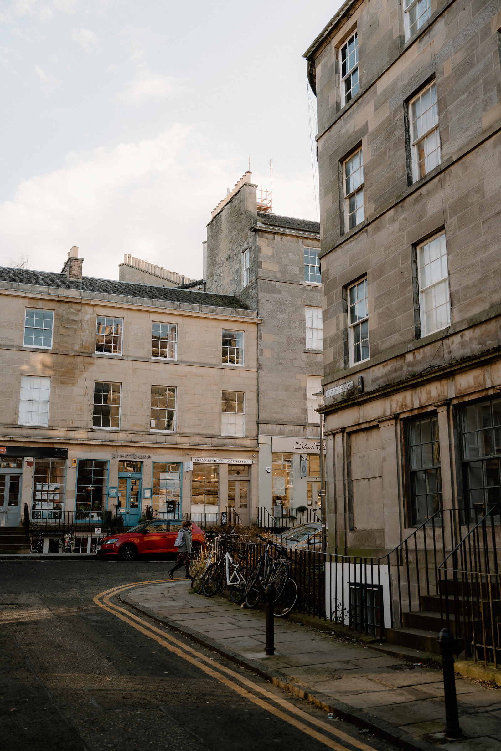 A red car is parked outside sandstone shopfronts on a curving Edinburgh street.