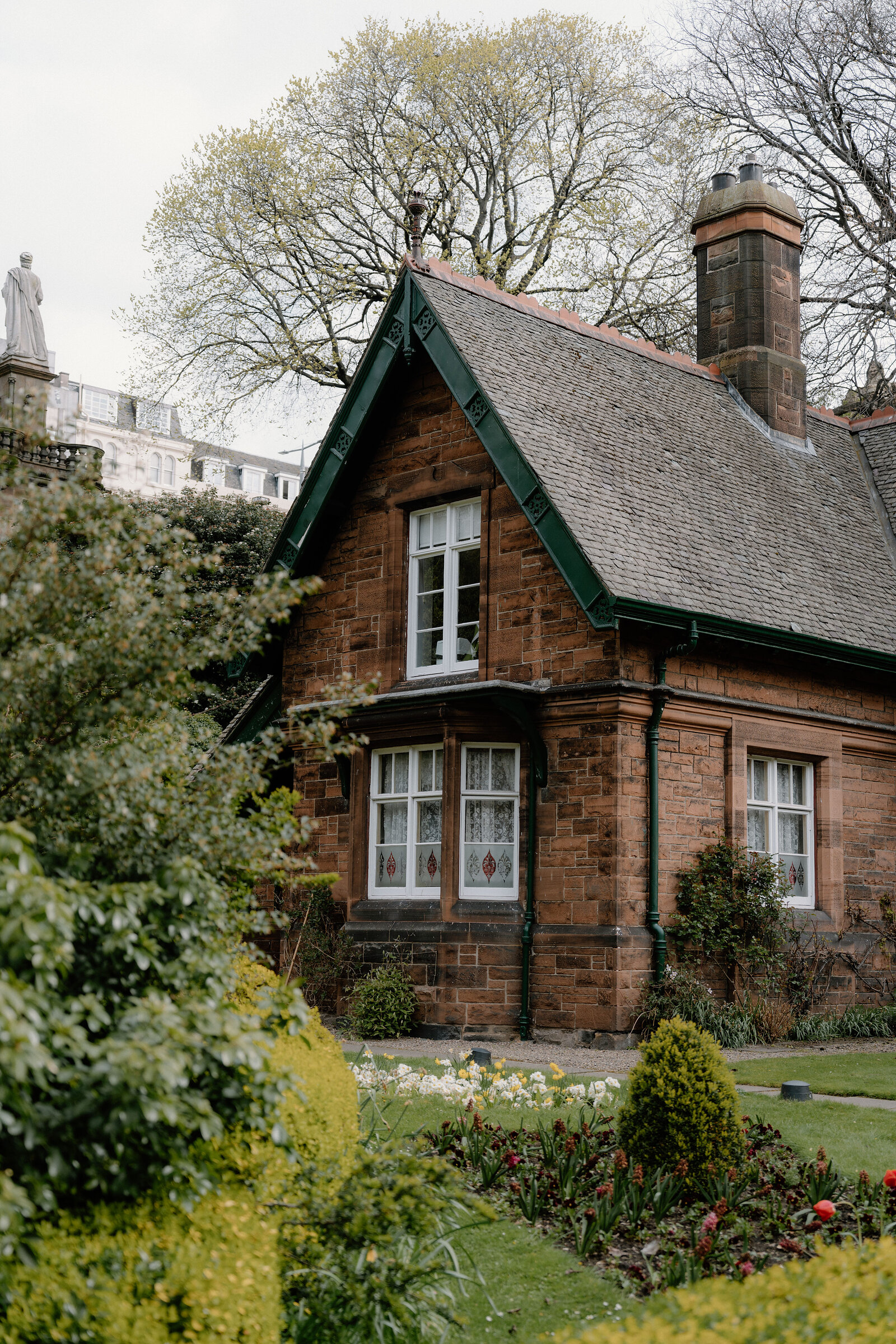 A red sandstone cottage with green-trimmed gables sits behind a hedge and spring flowerbeds, a statue visible beyond.
