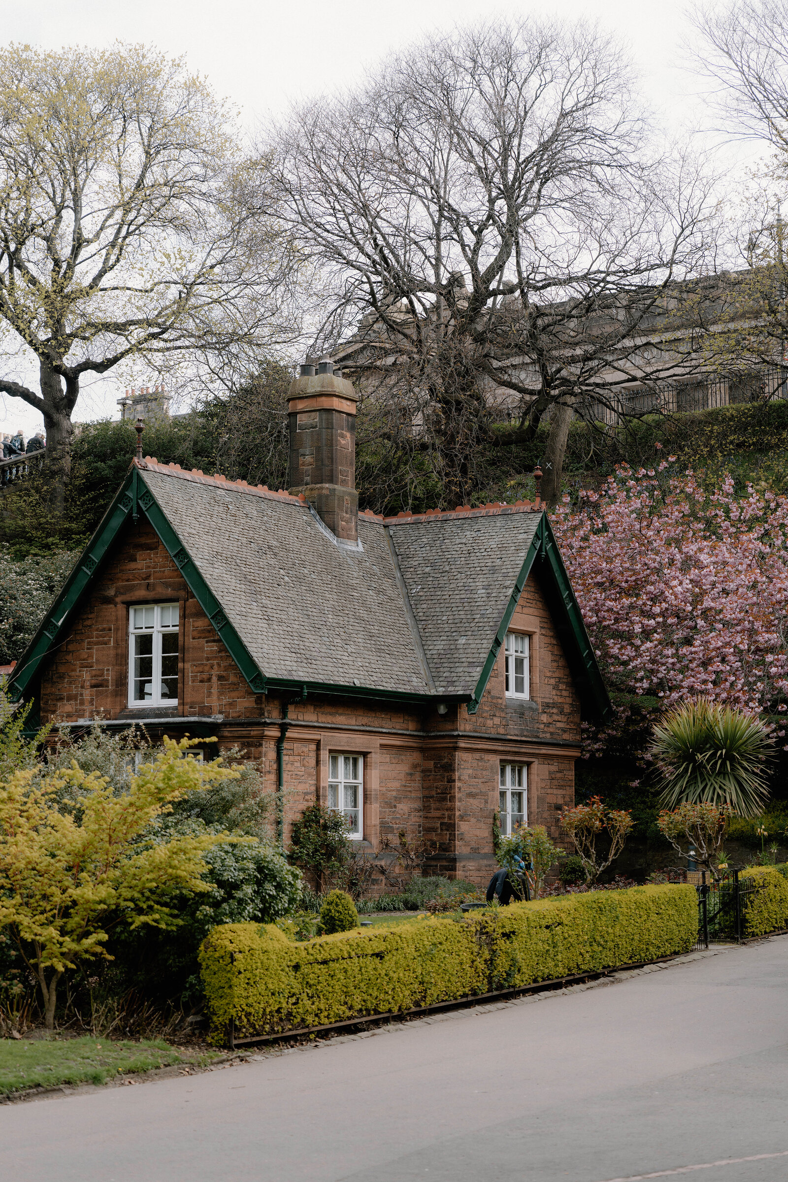 A red sandstone cottage with a tall chimney stands before blossoming trees and a stone building on the skyline.