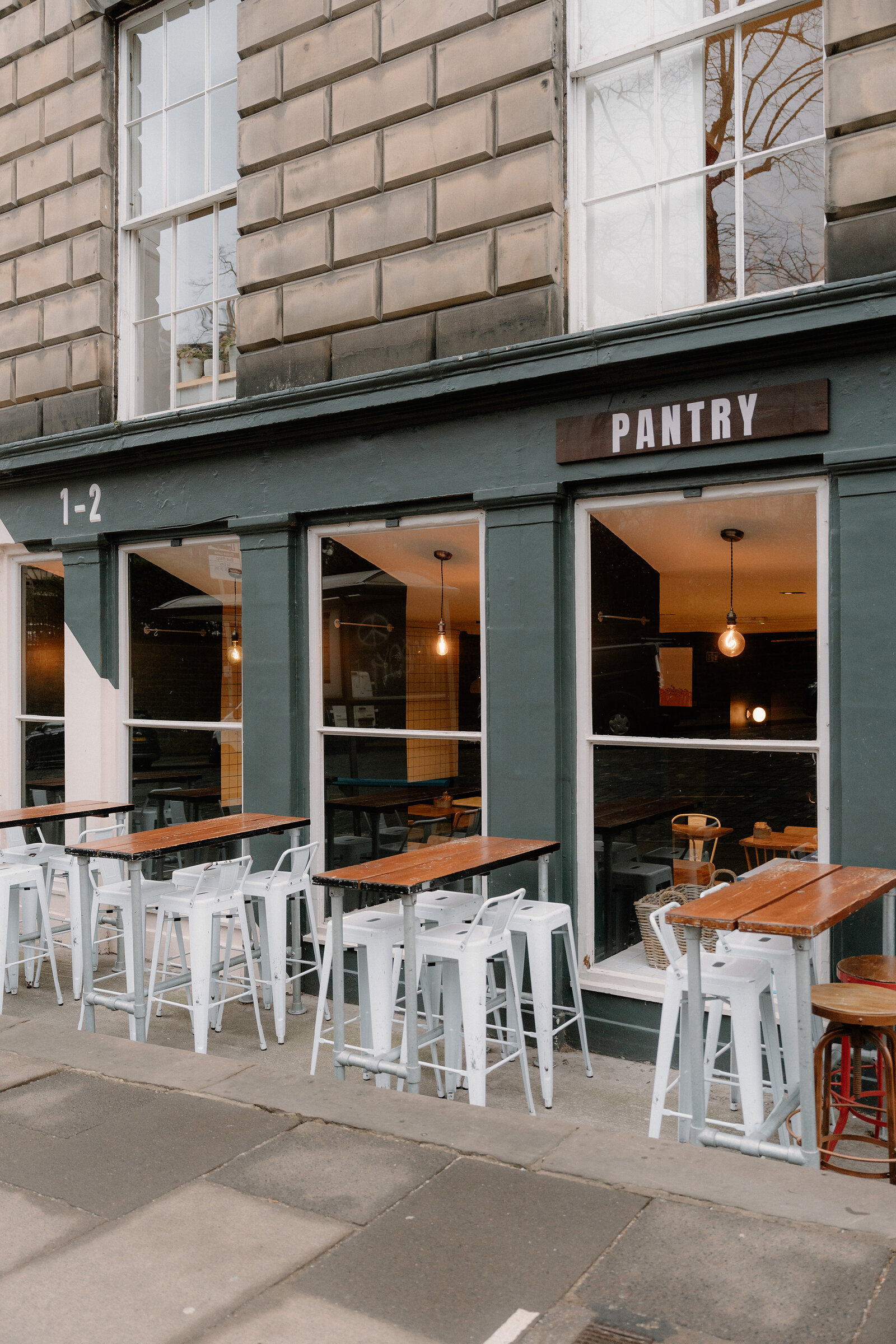 A sign reading 'Pantry' hangs above outdoor tables and stools on a green-painted Edinburgh shopfront.