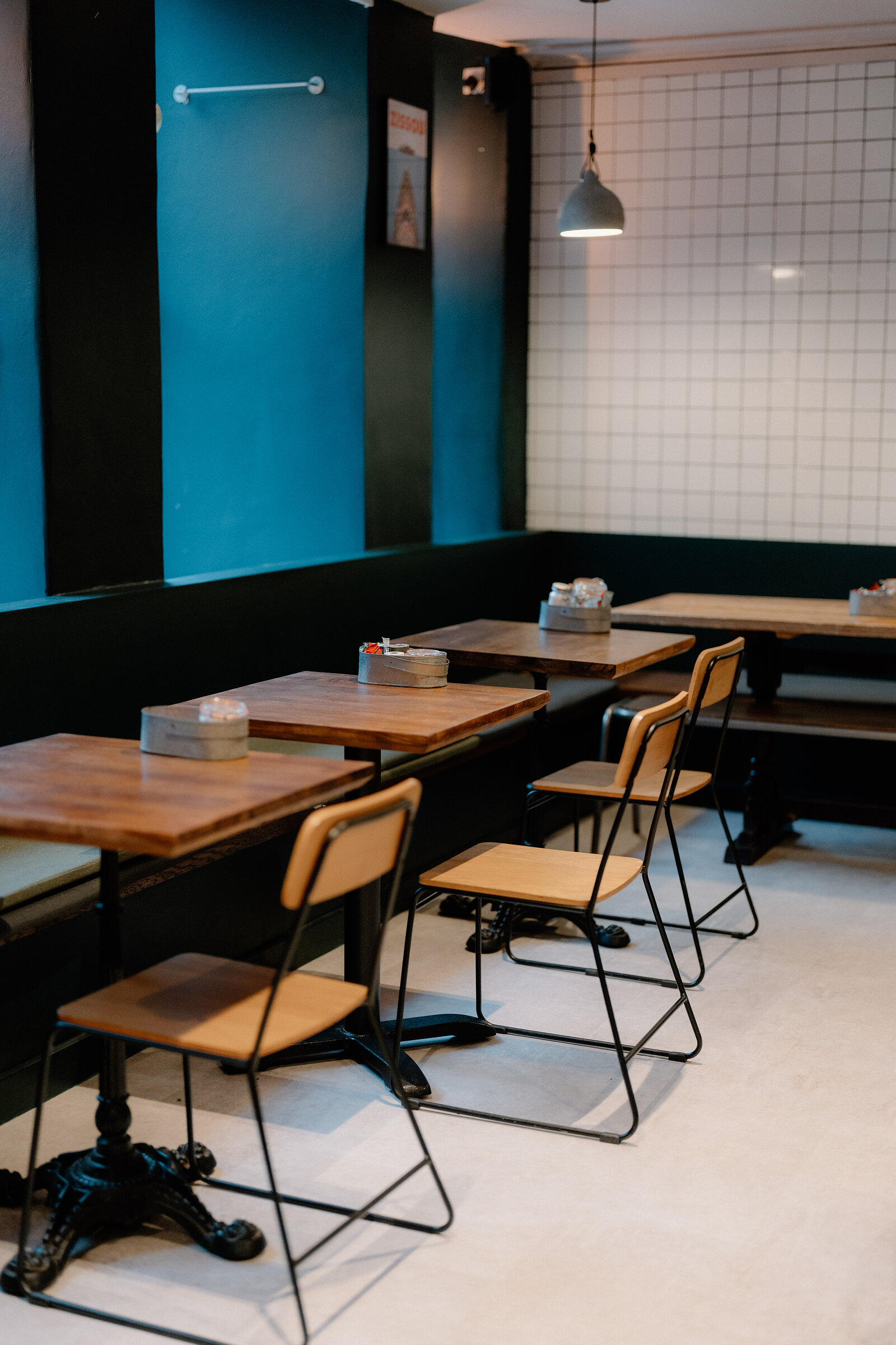 Wooden bistro tables and chairs line a teal and black wall beneath a white grid-tiled wall and pendant light.