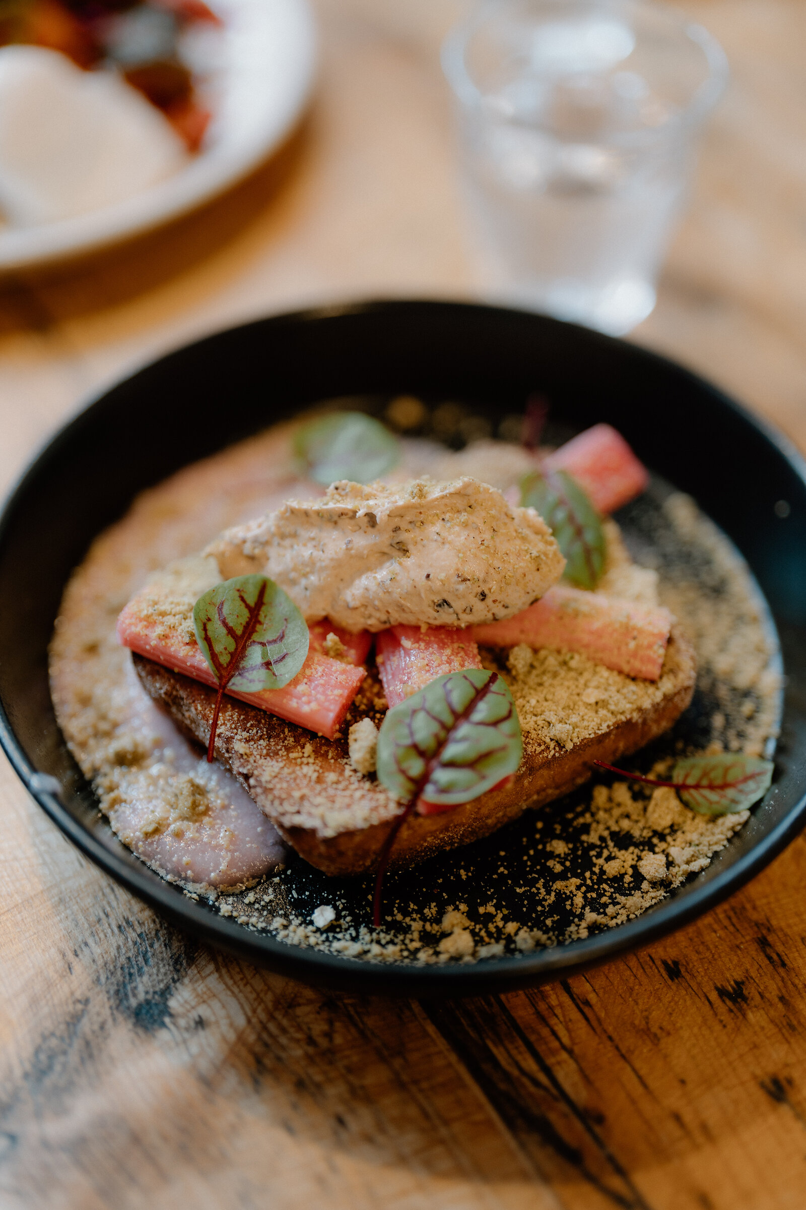 Toasted bread topped with rhubarb, sorrel leaves and crumble sits in a black bowl on a wooden table.
