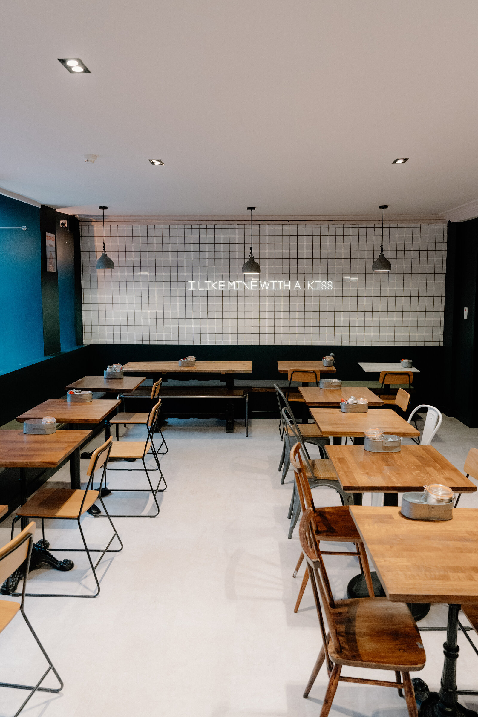 A neon sign reading 'I like mine with a kiss' glows above cafe tables and chairs in a tiled dining room.