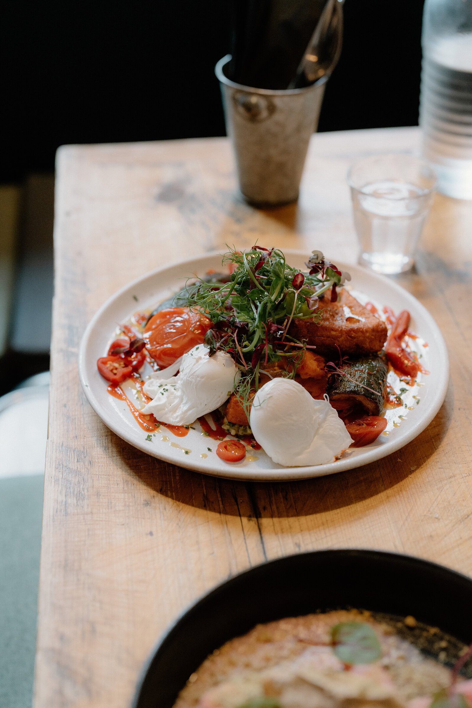 A plate of poached eggs, burrata and roasted tomatoes sits on a wooden table beside a metal ice bucket.