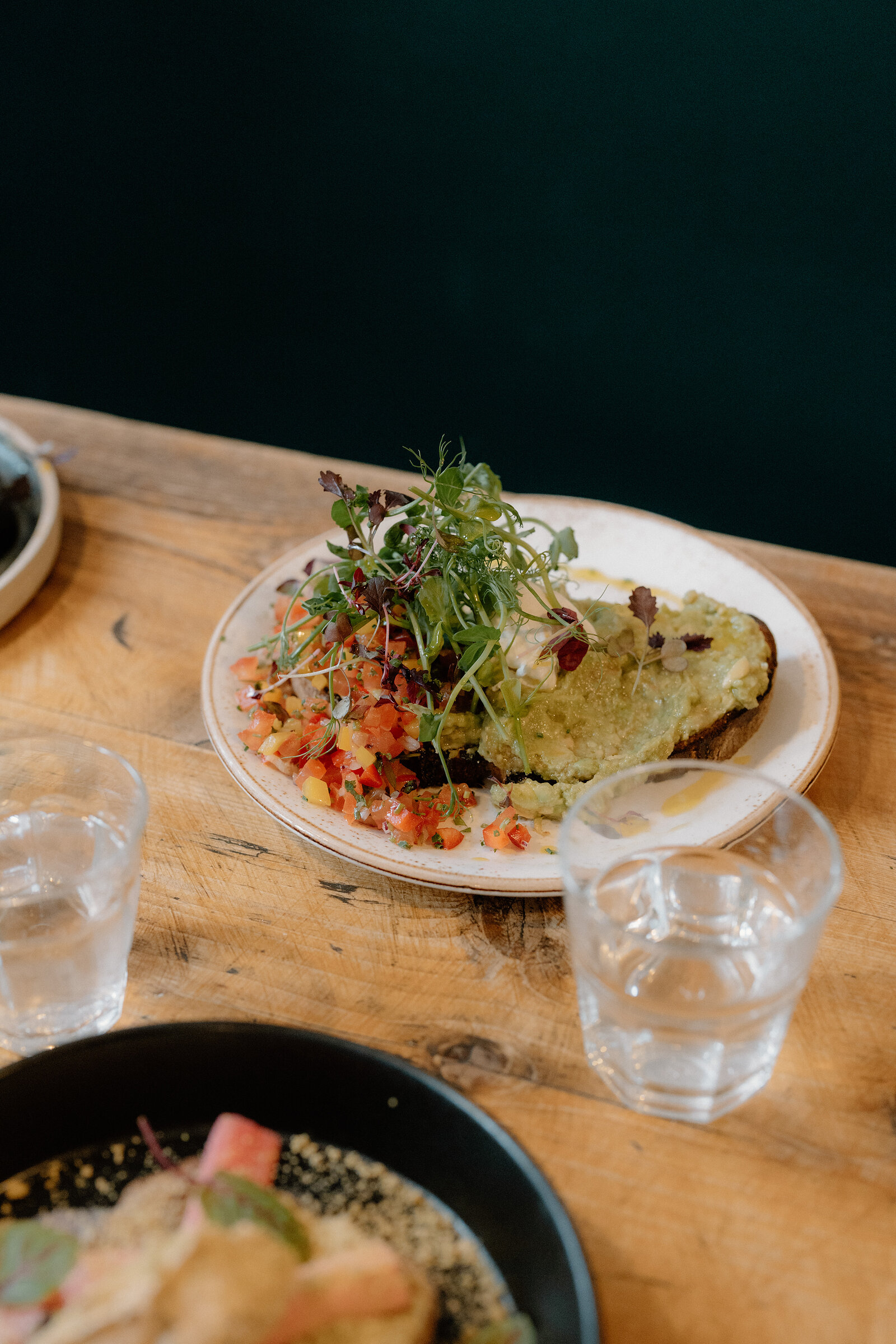 Avocado toast topped with microgreens and diced tomato sits on a plate beside two water glasses.