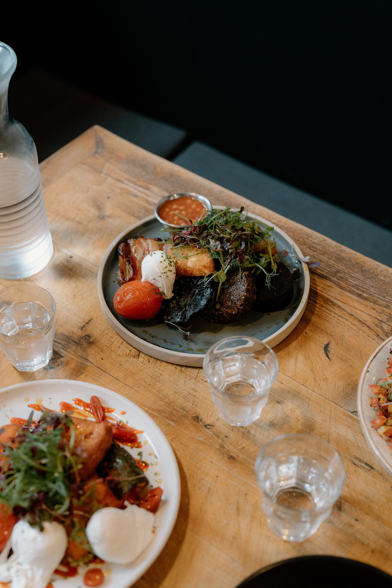 A plate of pork belly, poached egg, roasted tomato and greens sits on a wooden table with a water glass.