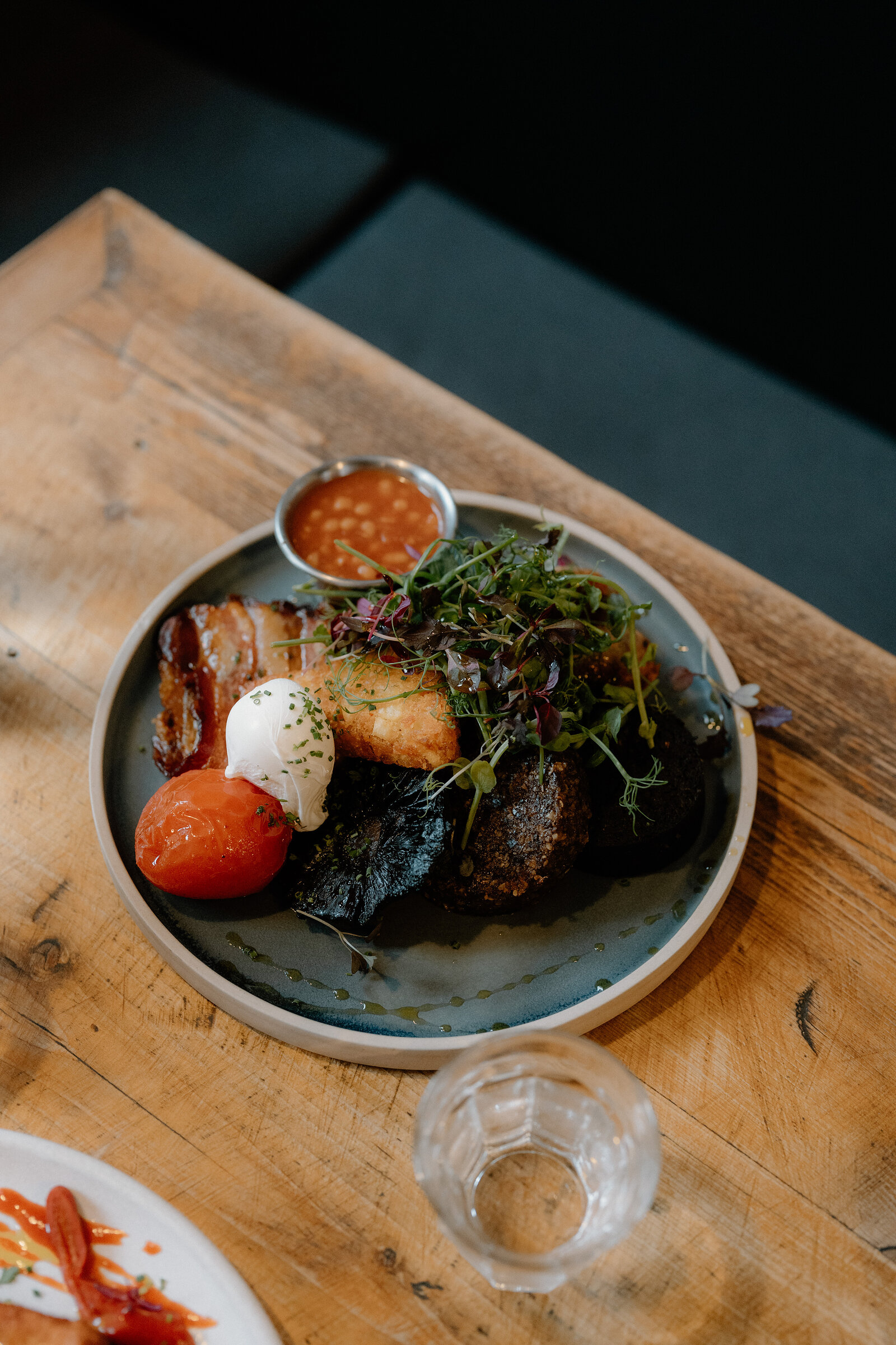 A plate of pork belly, poached egg and roasted tomato sits beside a small pot of sauce.