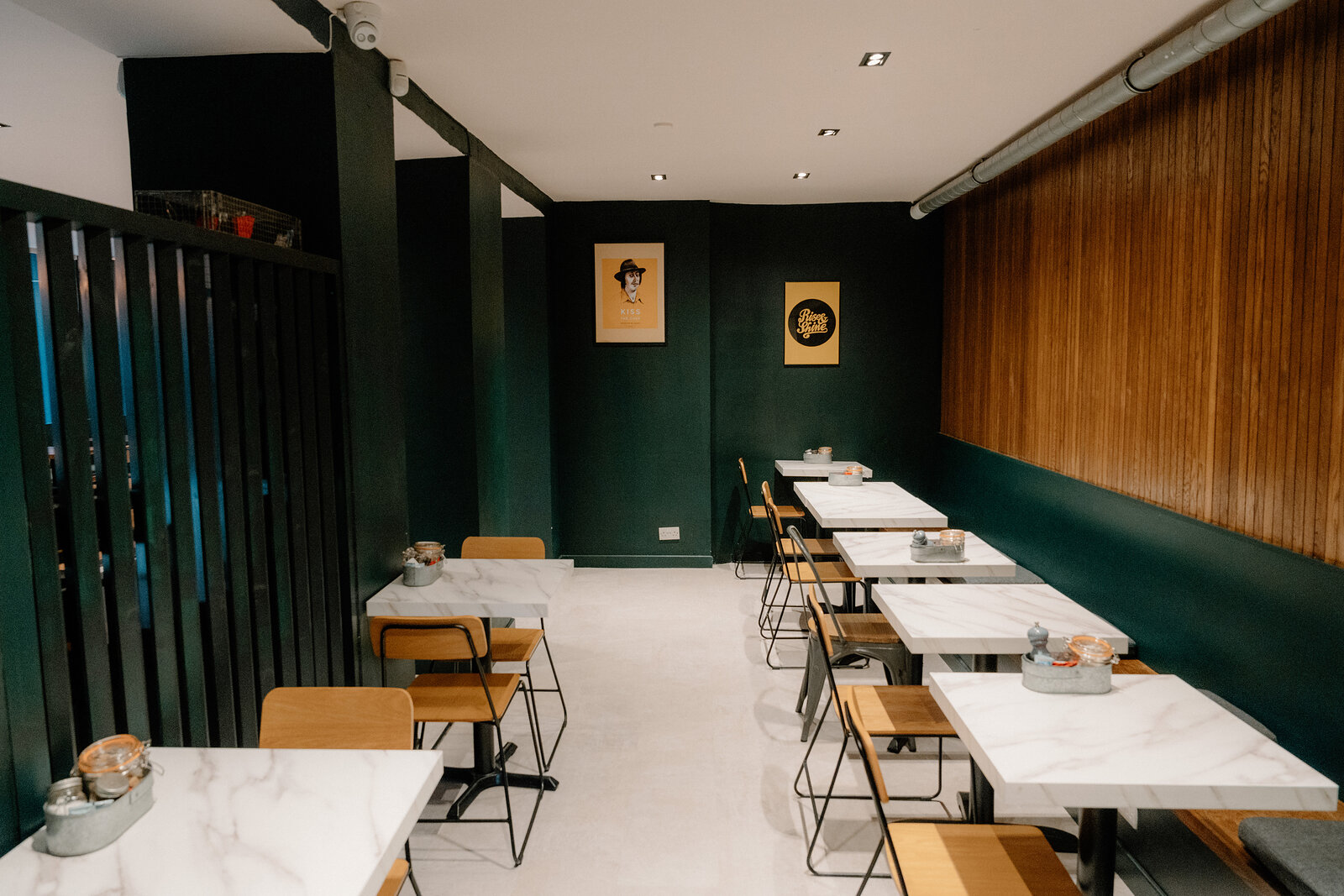 Framed illustrated posters hang on a dark green wall above marble tables in a narrow dining room.