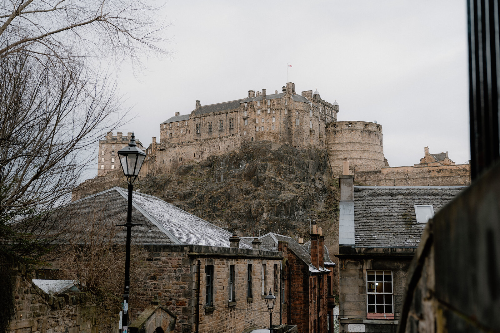 Edinburgh Castle rises above rooftops and bare trees, seen from below.