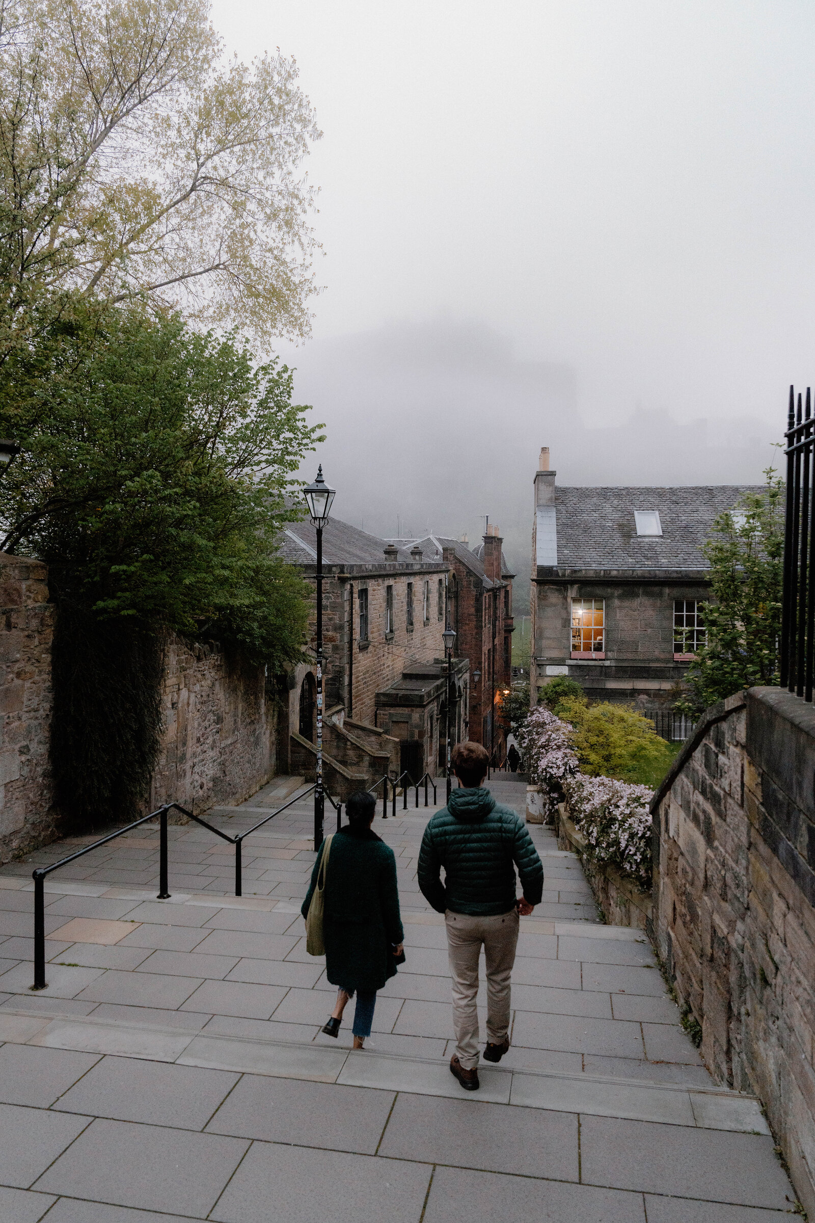 Two people walk down a misty stepped lane past stone buildings and blossoming trees.