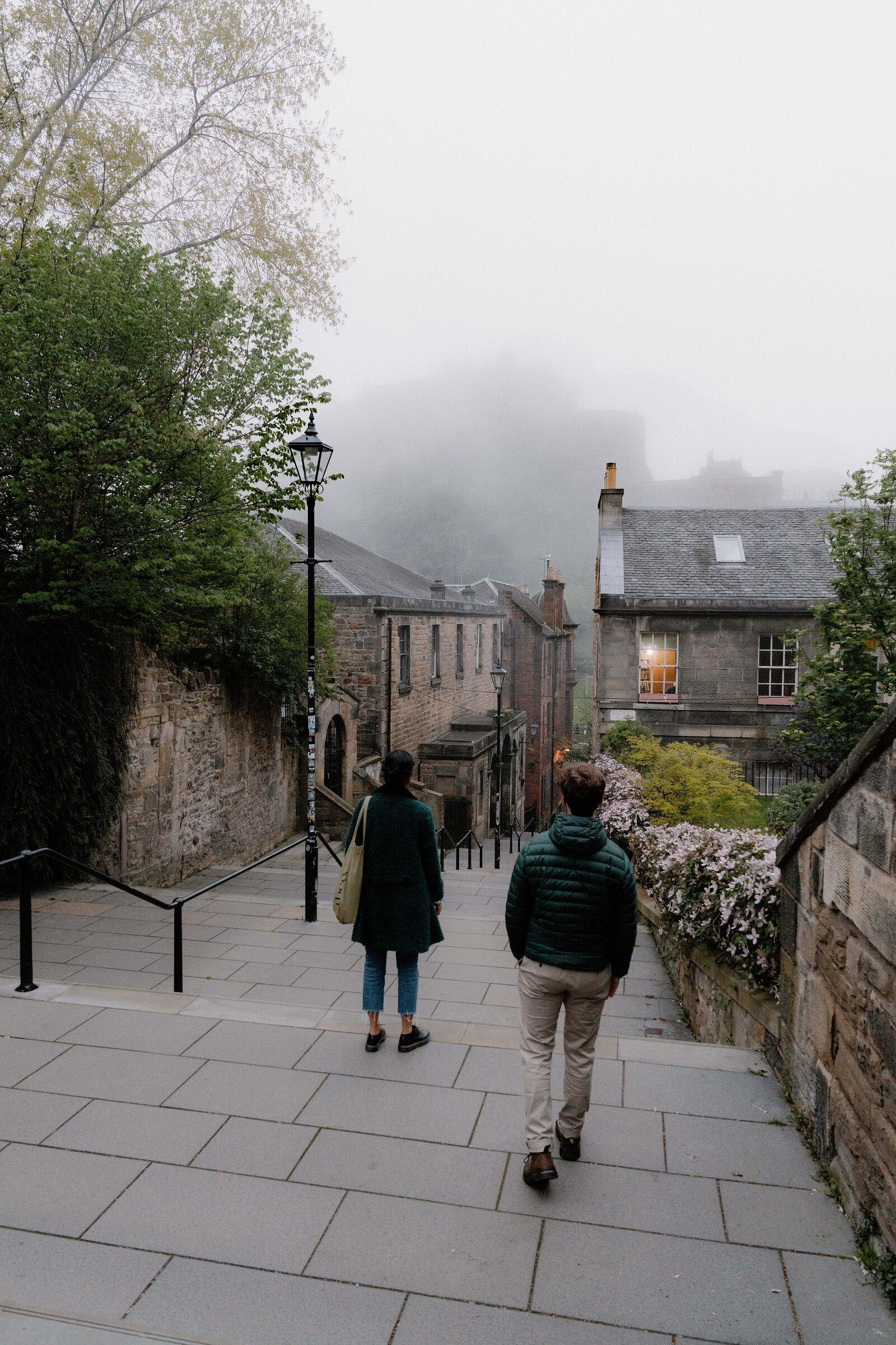 Two people walk down a misty stepped lane toward Edinburgh Castle, blossom trees along the wall.