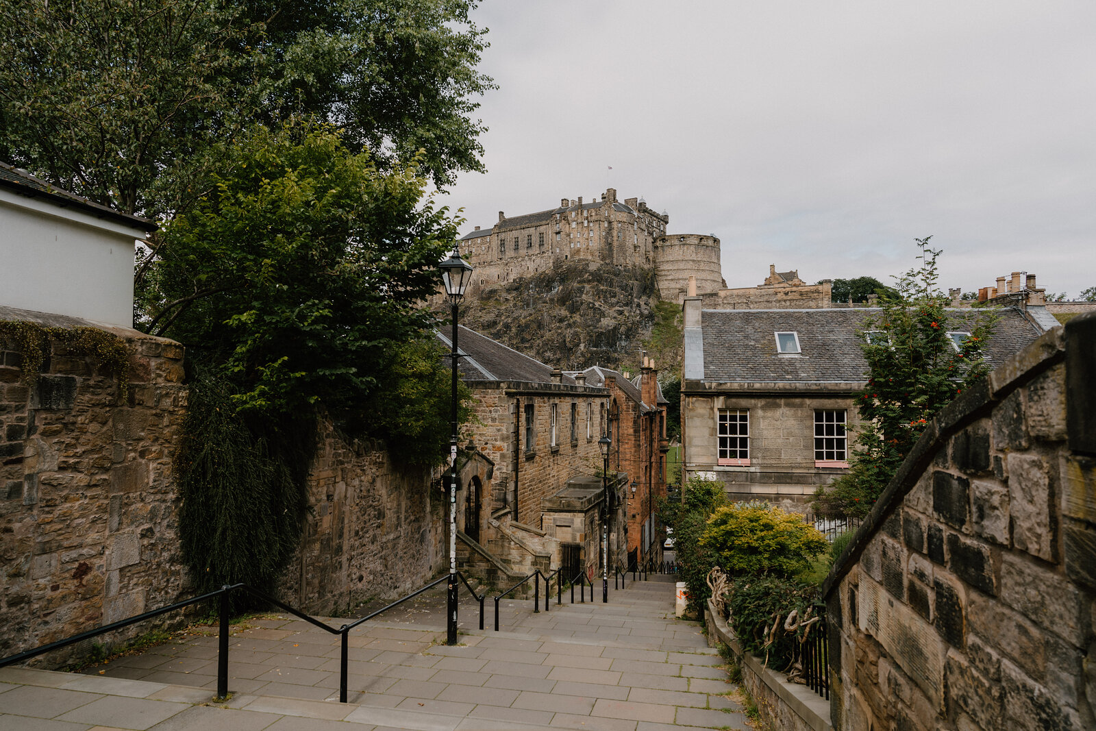 A lamppost and stone wall line a stepped lane descending toward Edinburgh Castle.