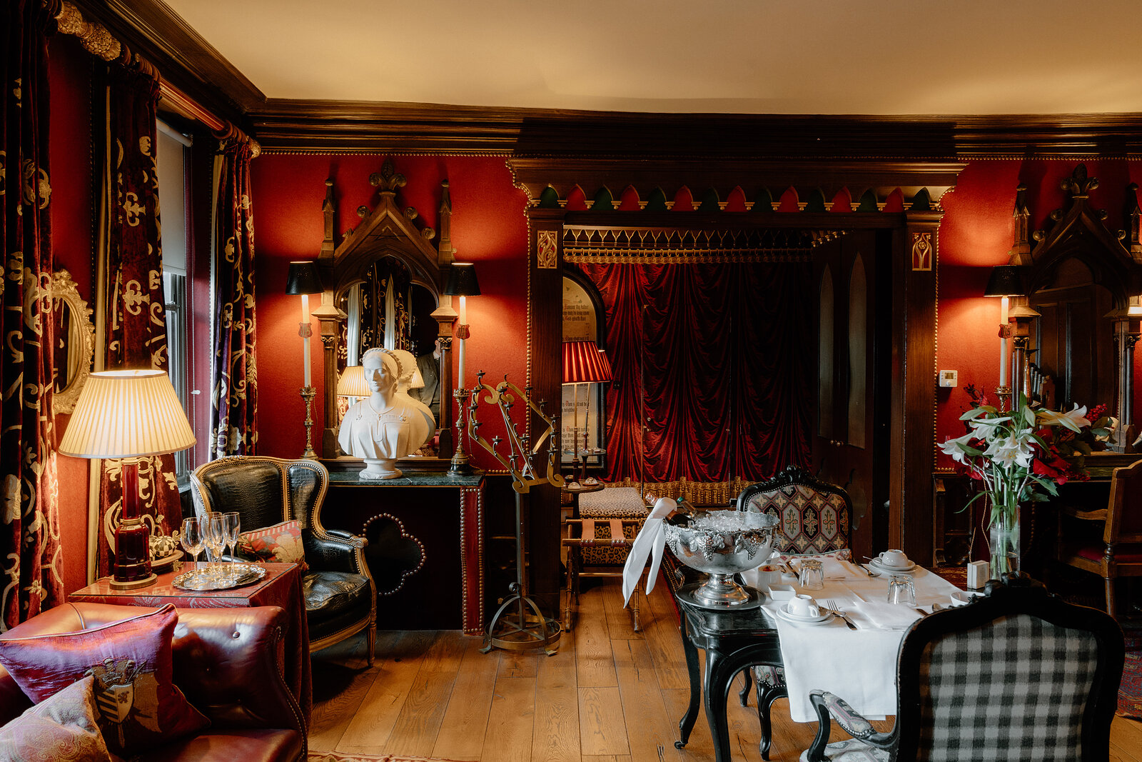 A carved statue and mirrored cabinet sit beside a table set for dining in a red-walled Gothic room.