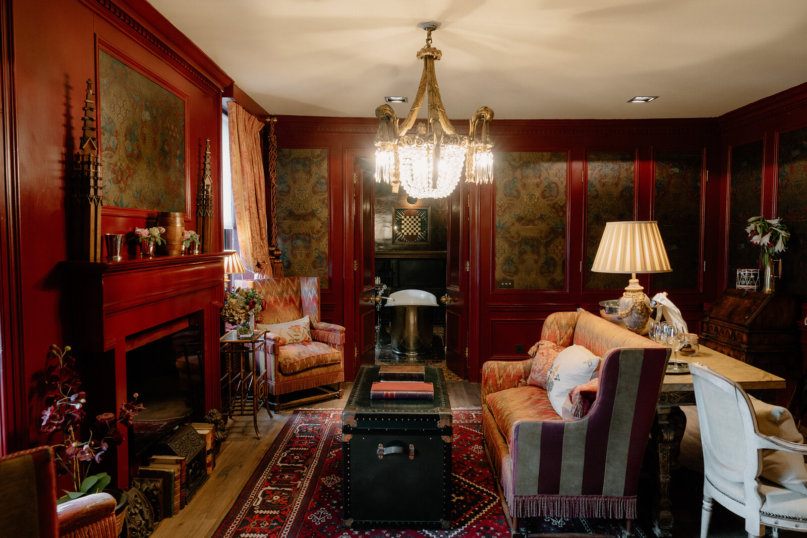 A red-panelled sitting room holds armchairs, a chandelier and a fireplace with a mirror above.