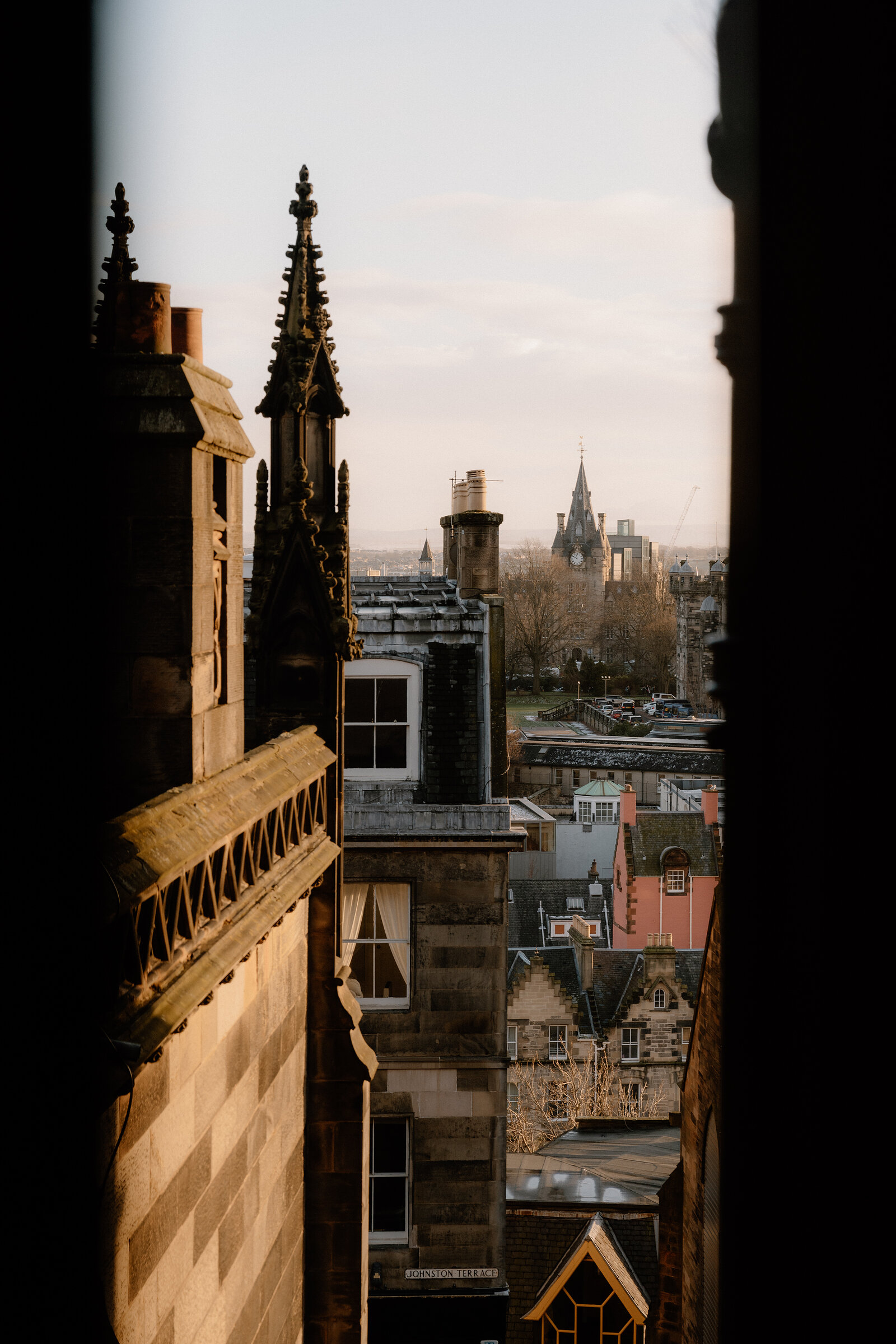 Spires and chimneys rise above Edinburgh's rooftops, seen through a window at golden hour.