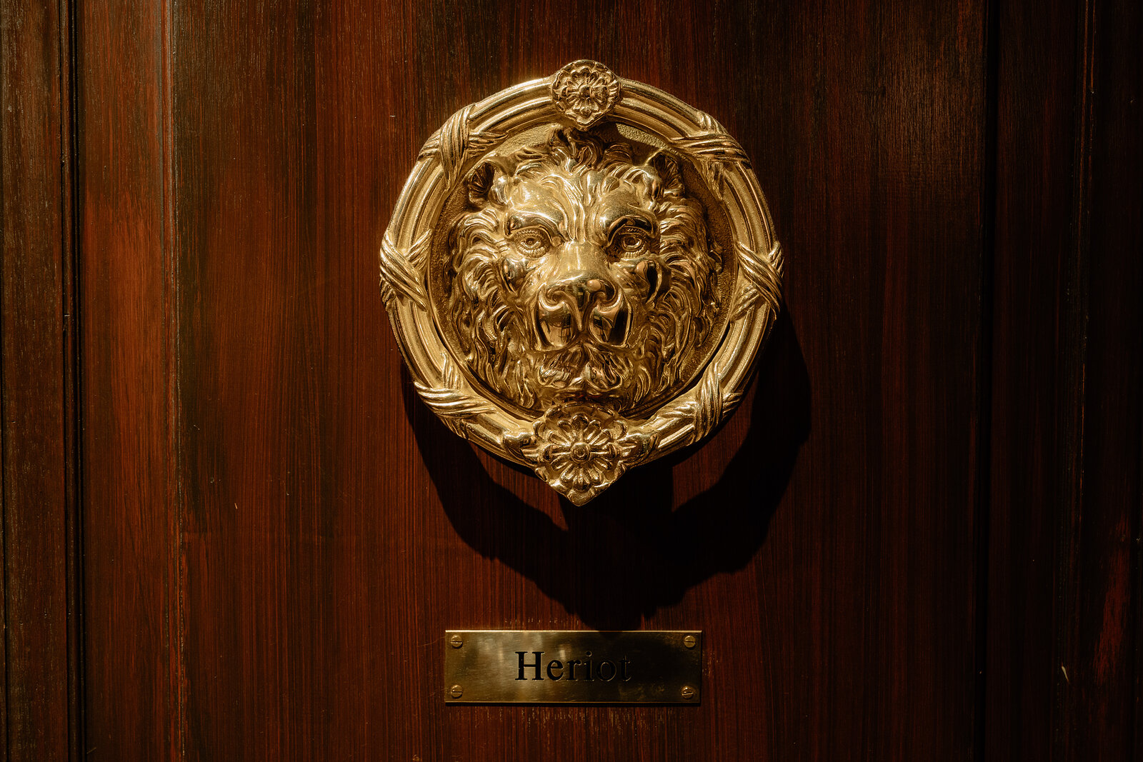 A brass lion's-head door knocker hangs above a nameplate reading 'Heriot' on a wooden door.