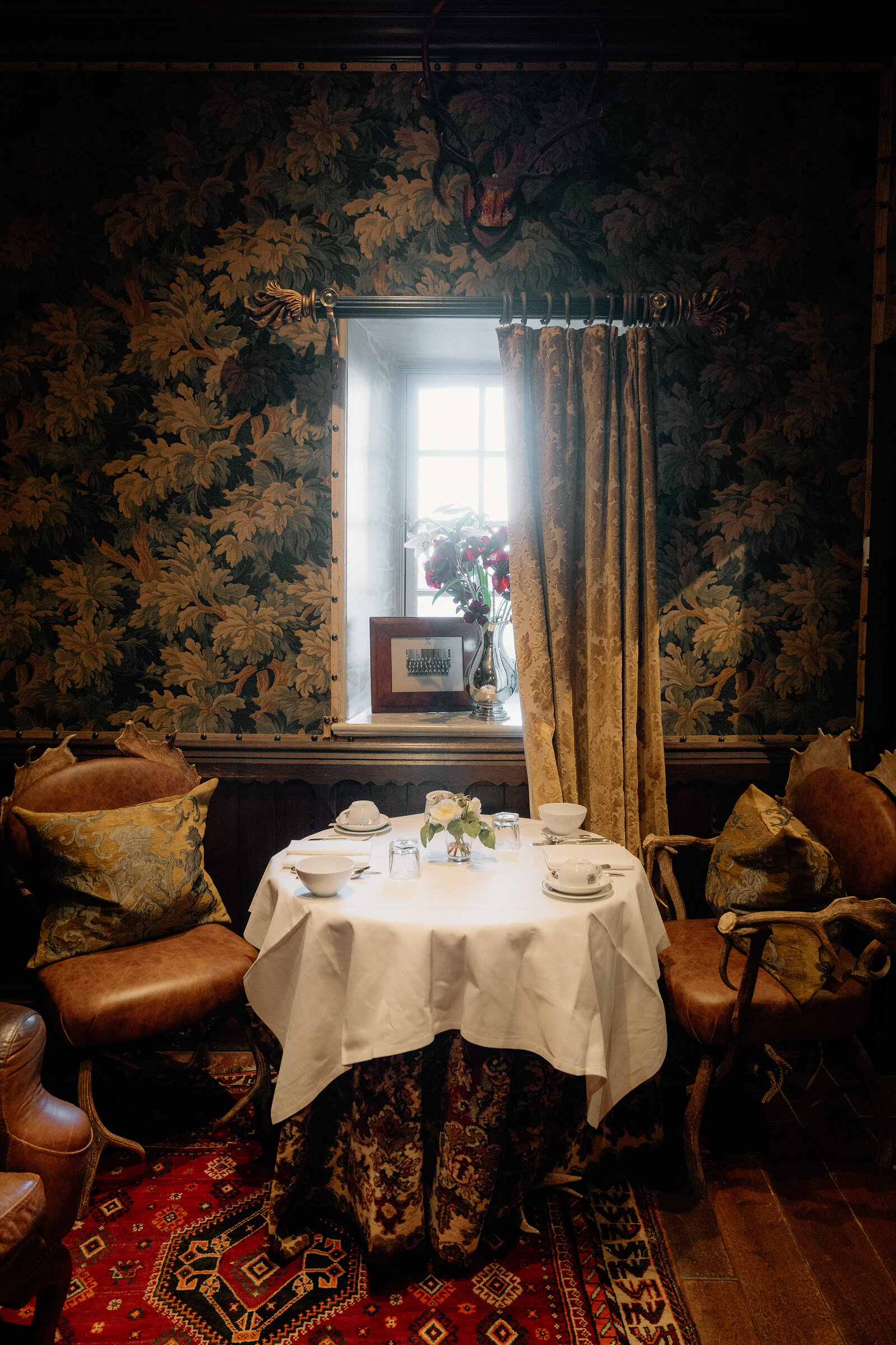 A round table set for dining sits before a curtained window in a tapestry-walled room.
