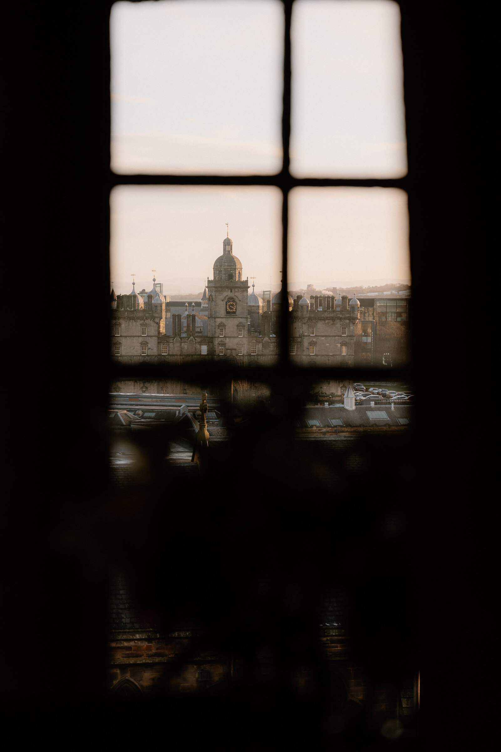 A domed clock tower rises above sandstone buildings, seen through a small window at dusk.