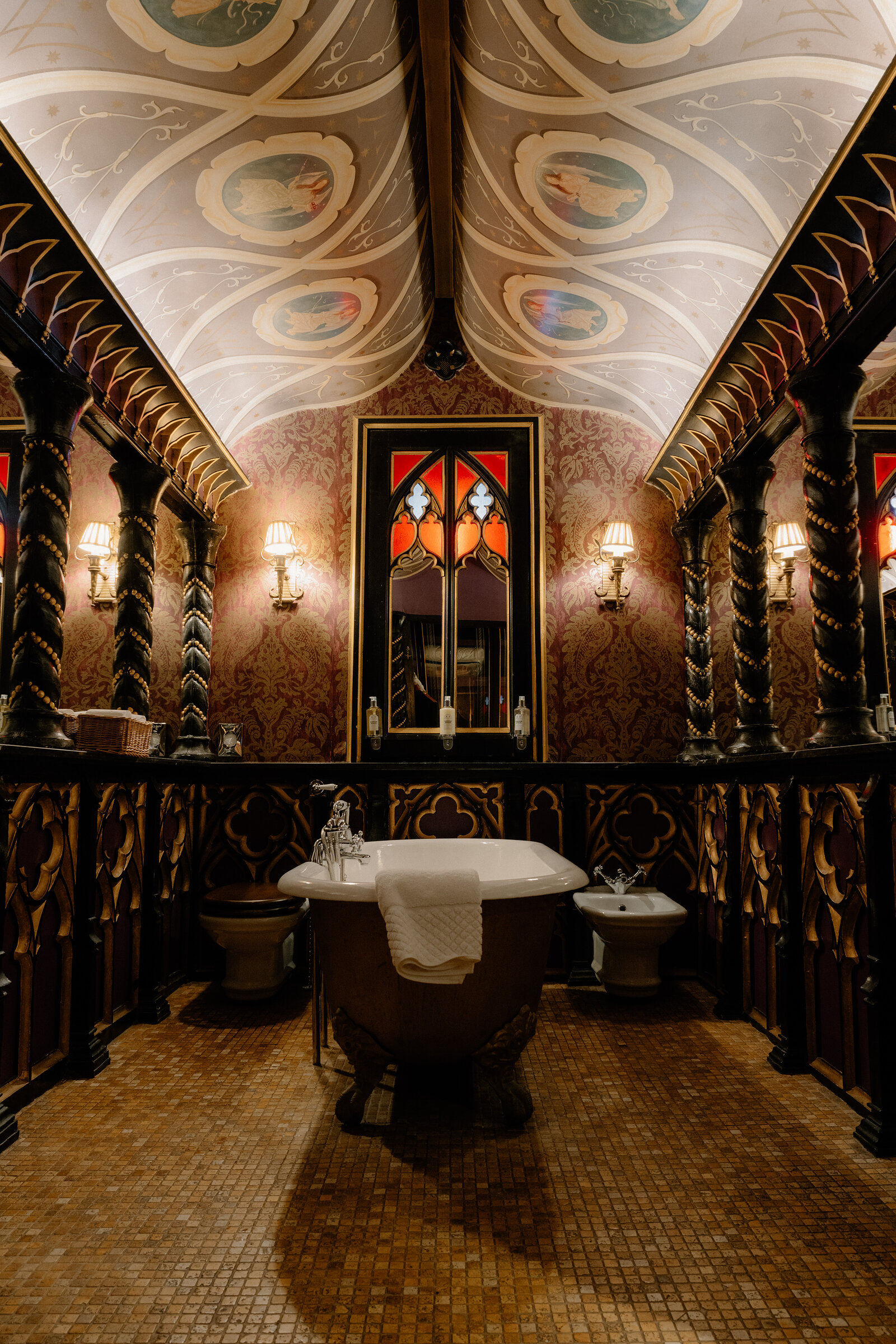 A painted vaulted ceiling arches above a clawfoot bath flanked by carved wooden pillars and stained glass.