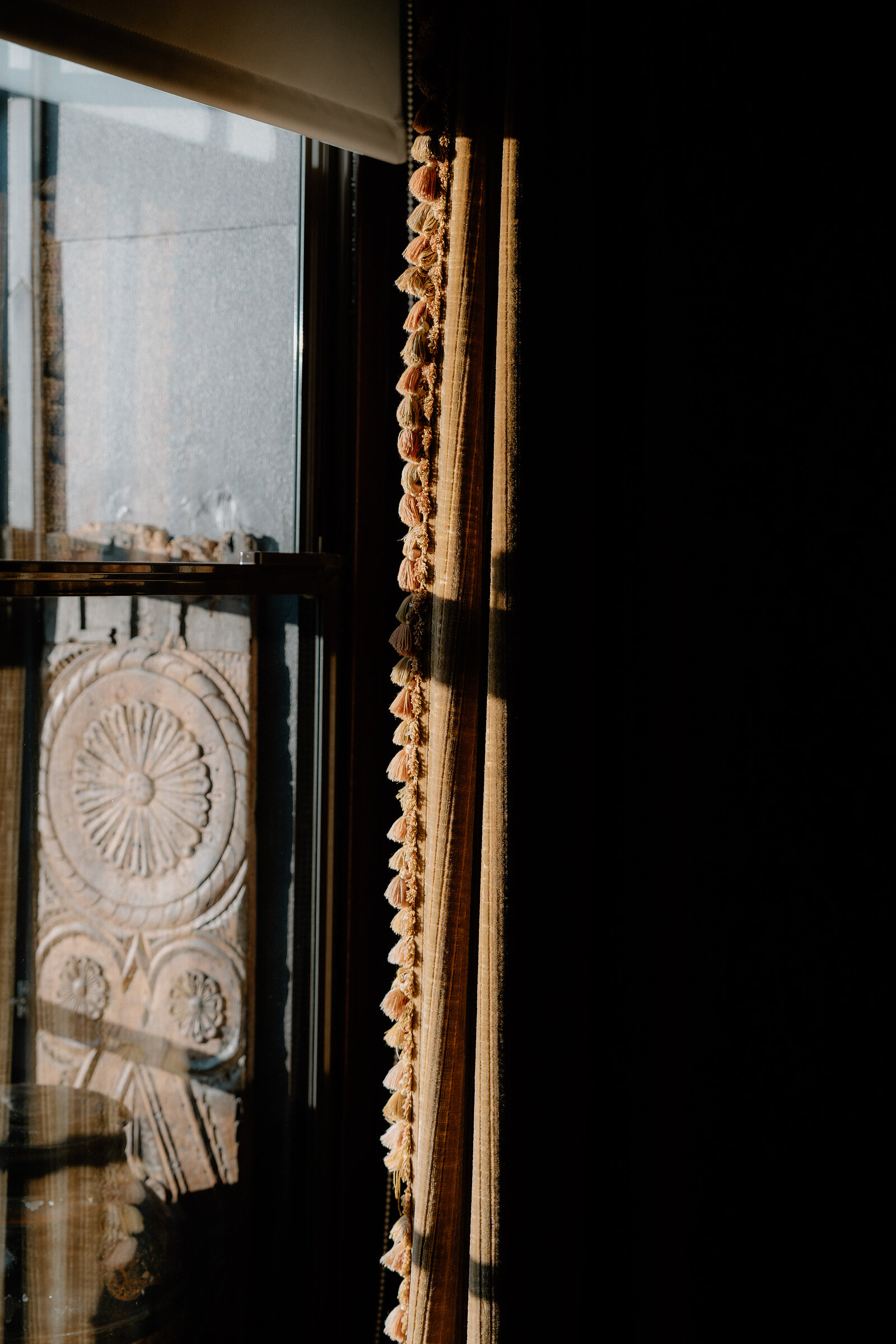 Sunlight catches a tasselled curtain beside a window reflecting a carved stone rosette.