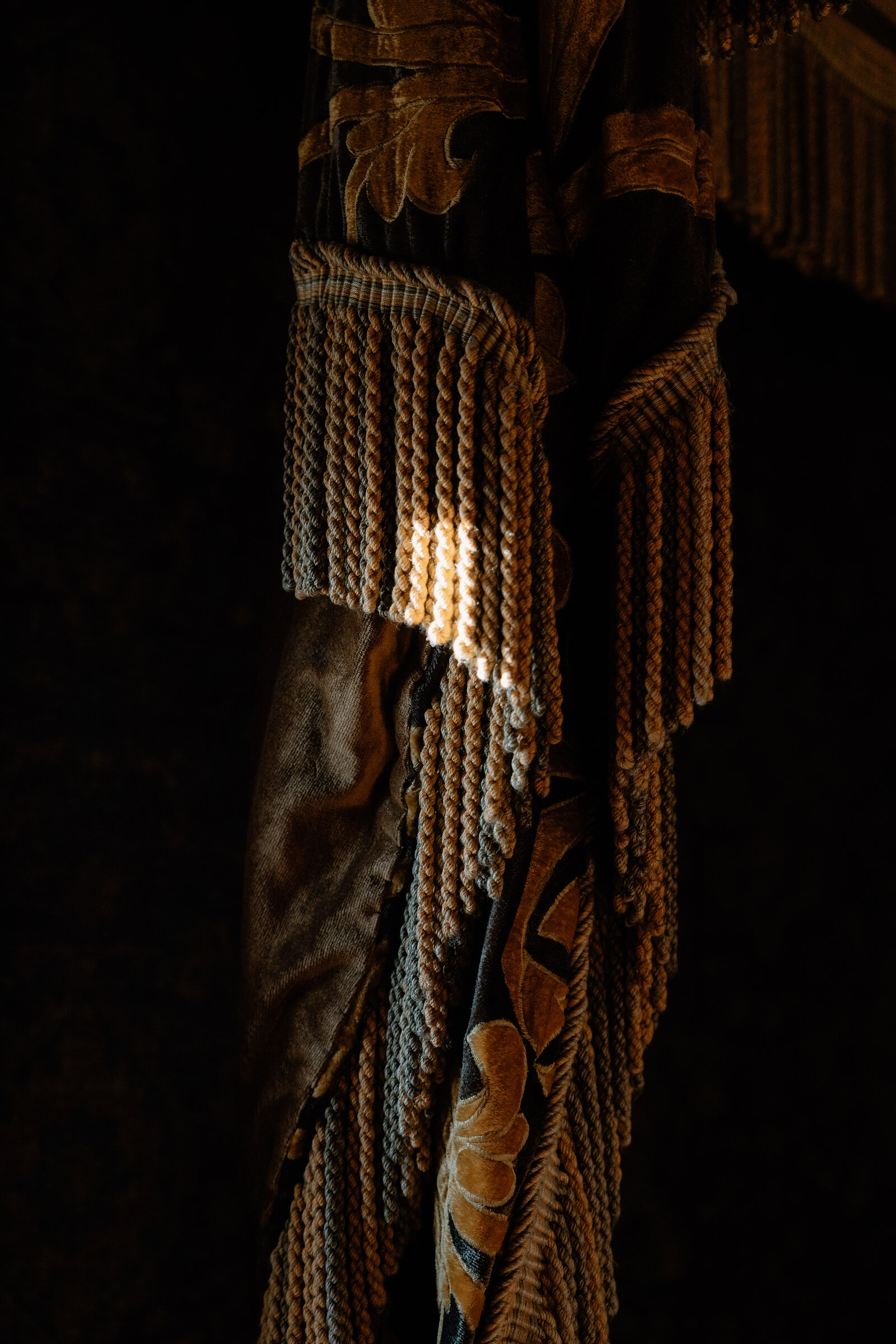 Sunlight catches the tasselled fringe of an ornate patterned curtain.