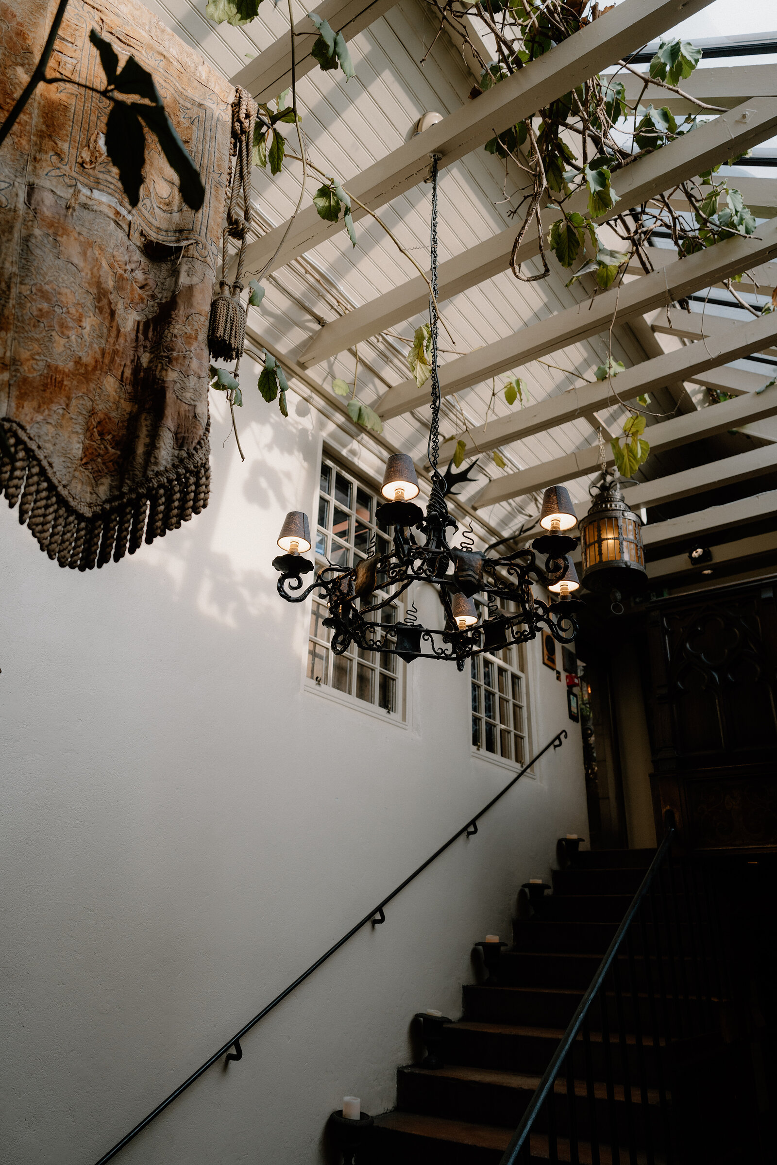 A weathered tapestry hangs above a stone staircase lit by a wrought-iron chandelier beneath a pergola.
