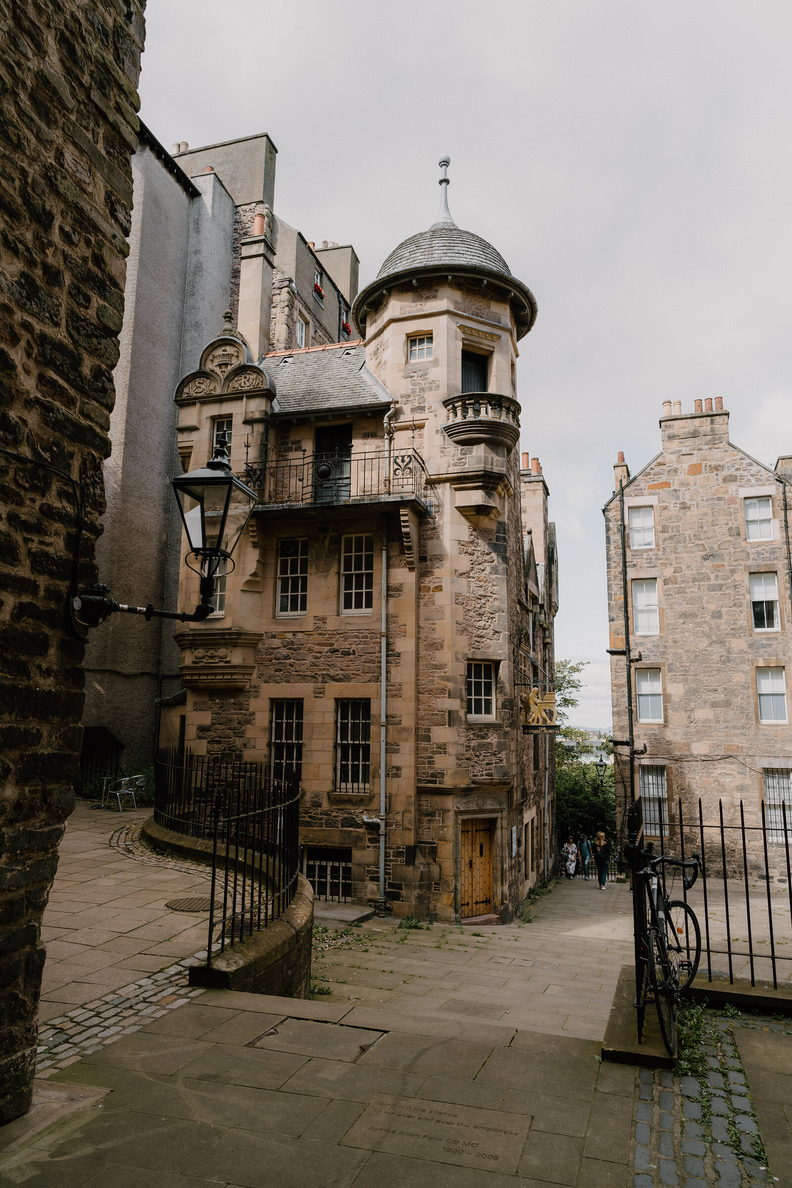 A turreted sandstone building with a corner tower rises above a cobbled close.