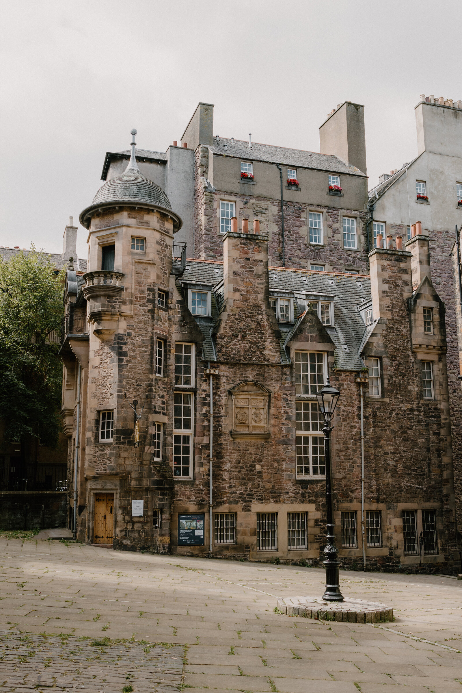 A turreted stone building with crow-stepped gables stands beside a lamppost on a paved square.