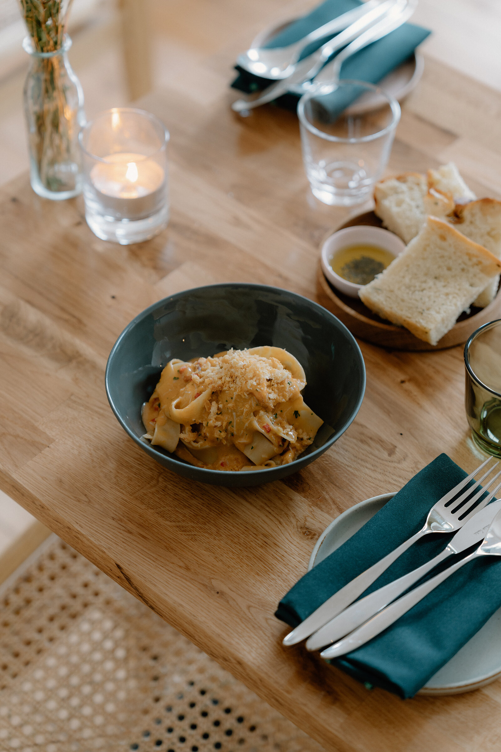 A dark bowl of pappardelle pasta topped with grated cheese sits on a wooden table beside sliced focaccia, olive oil, a lit tealight candle, and cutlery on a teal napkin.